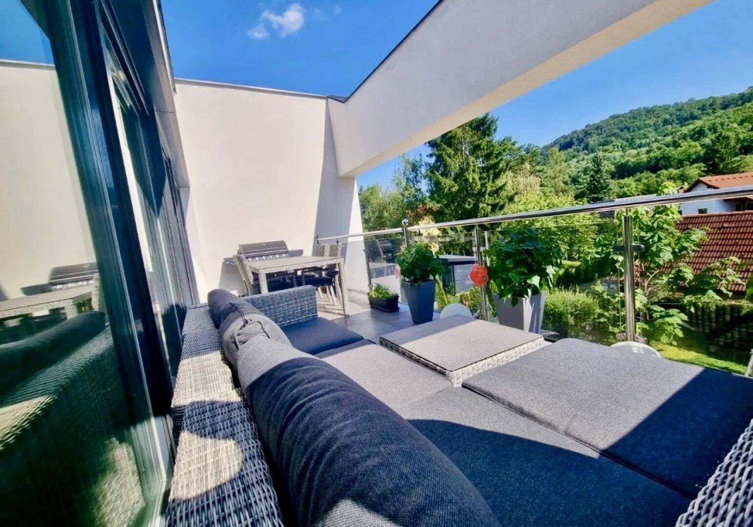 Sonnige Terrasse mit bequemen Loungemöbeln und Grillbereich, umgeben von grüner Landschaft.