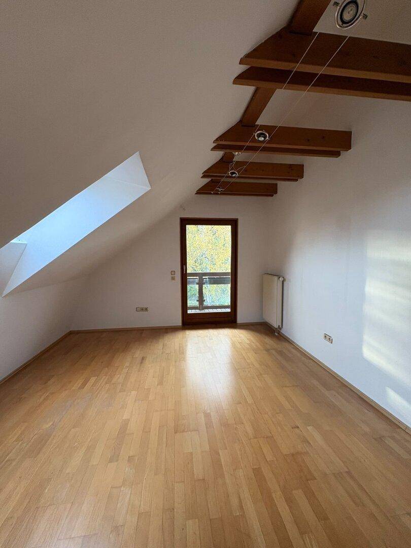 Heller Raum im Dachgeschoss mit Holzboden, sichtbaren Deckenbalken und Zugang zu einem kleinen Balkon.