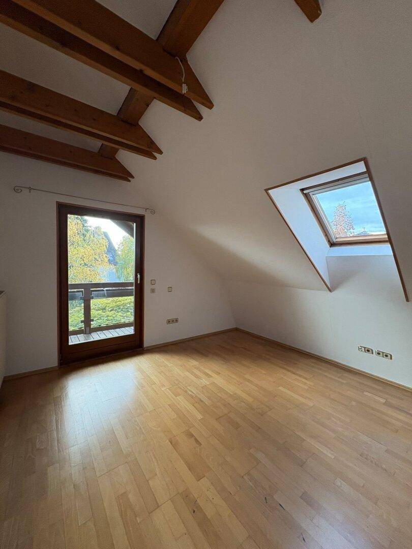 Lichtdurchfluteter Dachgeschossraum mit Holzboden, sichtbaren Balken, Dachfenster und Balkonzugang.