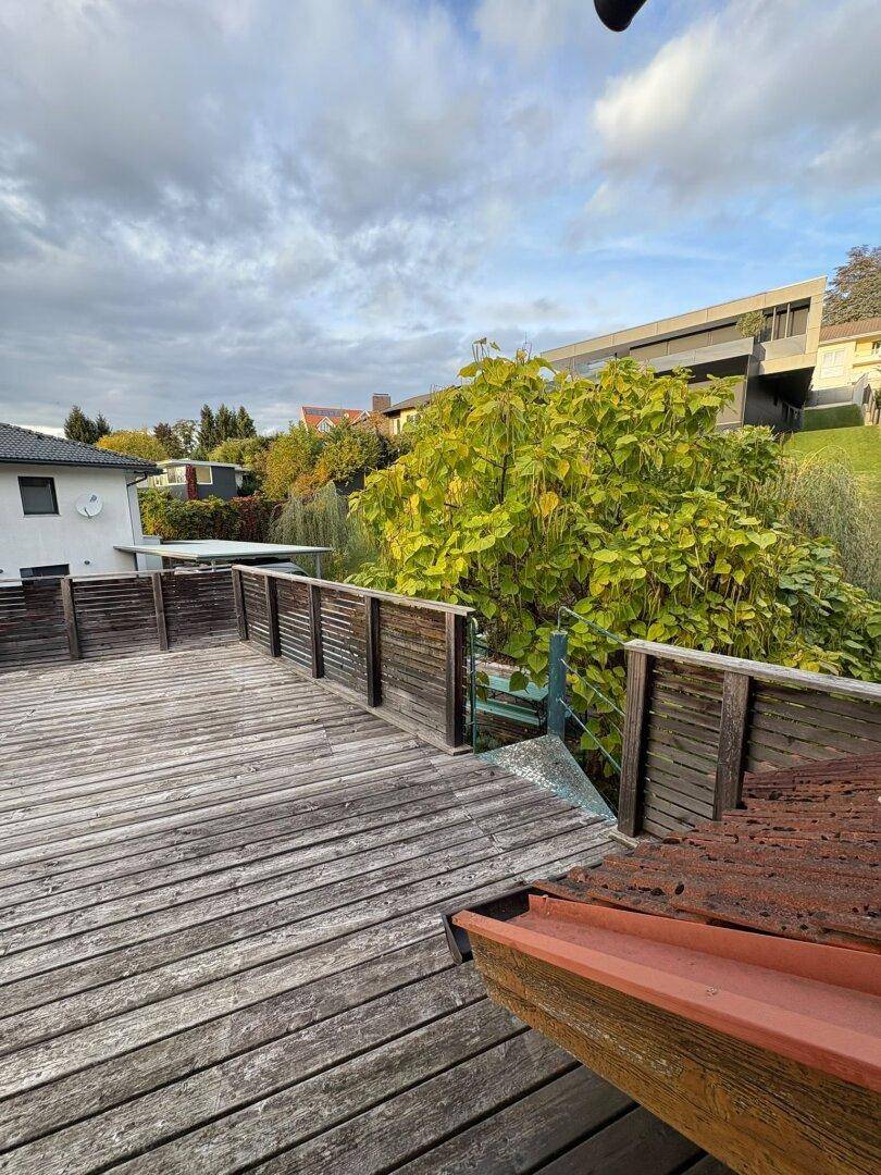 Geräumige Holzterrasse mit Blick auf moderne Architektur und üppiges Grün unter bewölktem Himmel.
