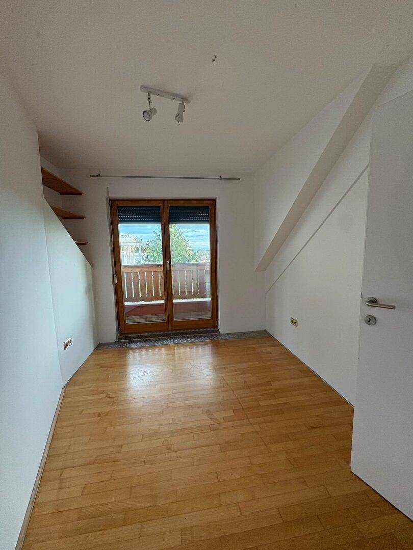 Leerer Raum im Dachgeschoss mit Holzboden, Dachschräge und Balkonzugang mit Rollläden.