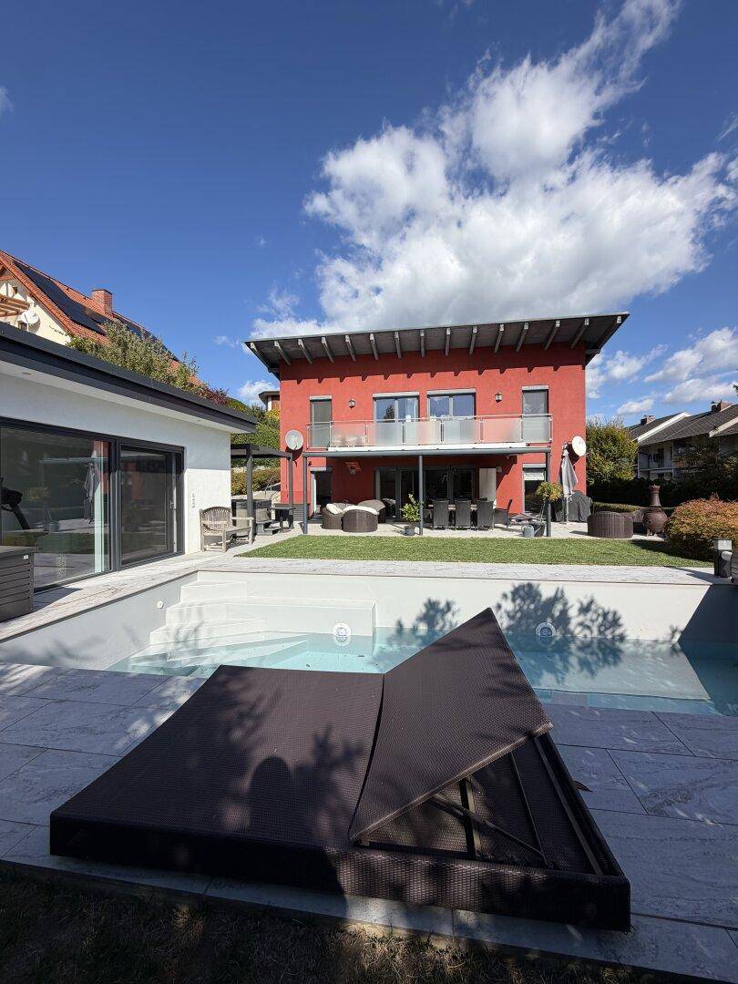 Rotes Wohnhaus mit modernem Poolbereich und Liegefläche unter strahlend blauem Himmel.