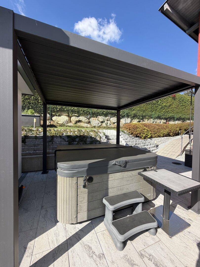 Überdachter Whirlpoolbereich auf der Terrasse, umgeben von einer gepflegten Hecke und Natursteinmauern.