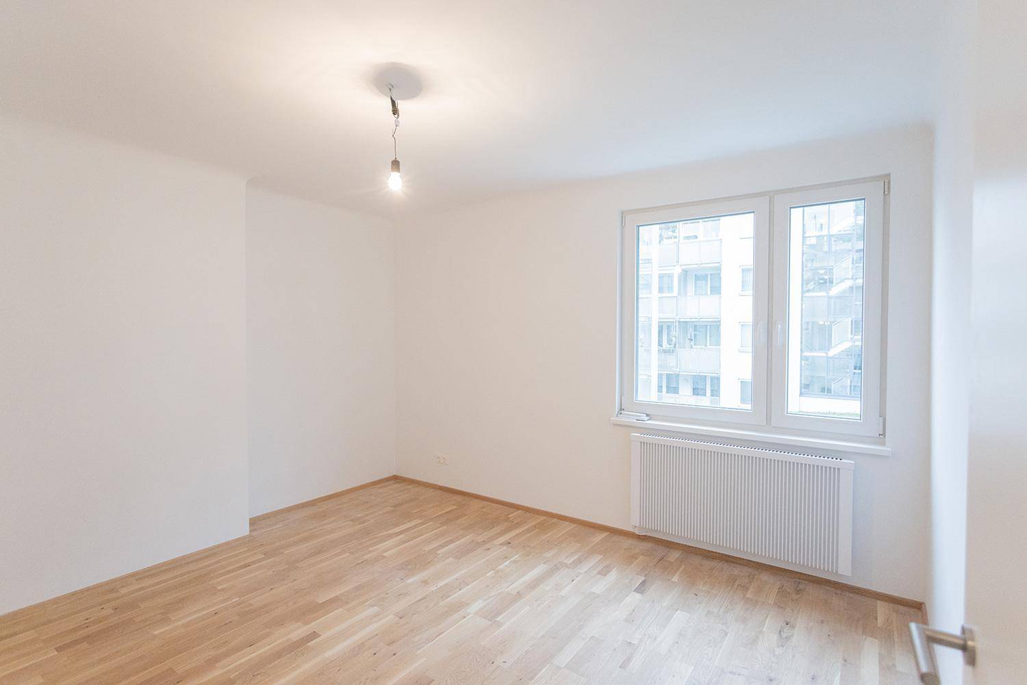 Helles, leeres Zimmer mit Parkettboden, großem Fenster und Heizkörper, bereit zur individuellen Gestaltung.