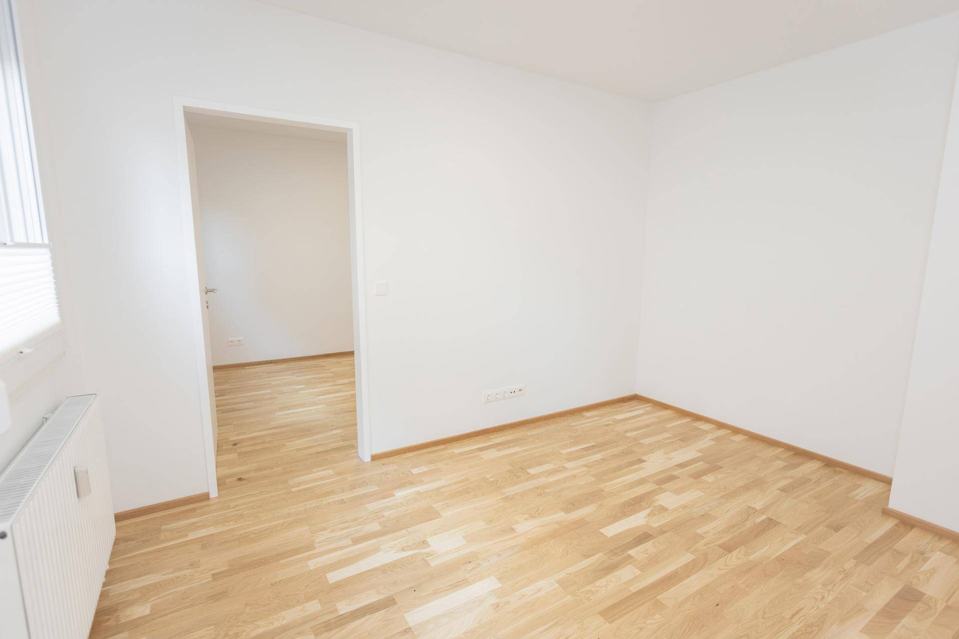 Heller, leerer Raum mit Parkettboden und weiß gestrichenen Wänden, mit Blick in einen weiteren Raum.