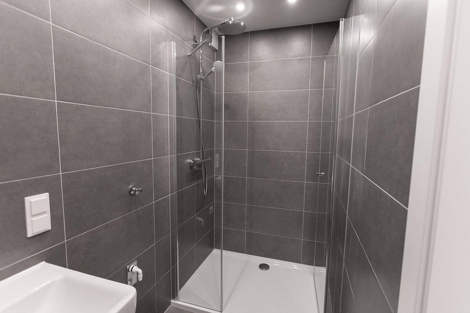 Modernes Badezimmer mit dunklen Fliesen, Duschkabine und kleinem Waschbecken.