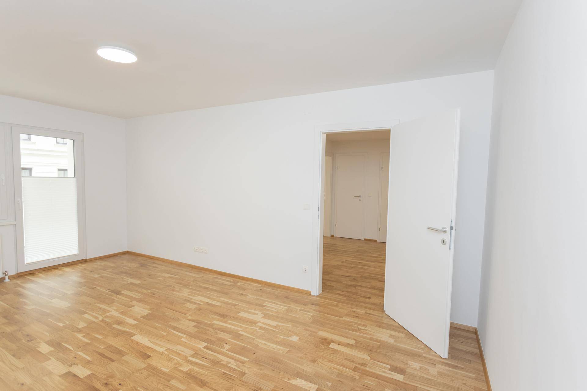 Heller Raum mit Parkettboden und offener Tür, die zu einem weiteren Zimmer führt.