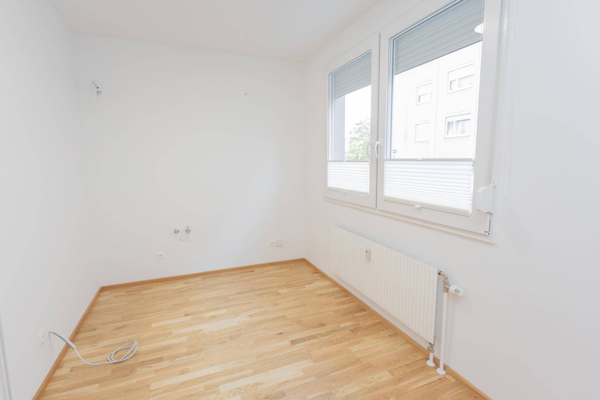 Leerer Raum mit hellem Parkettboden, Fenster mit Rollläden und Heizkörper, bereit zur Einrichtung.
