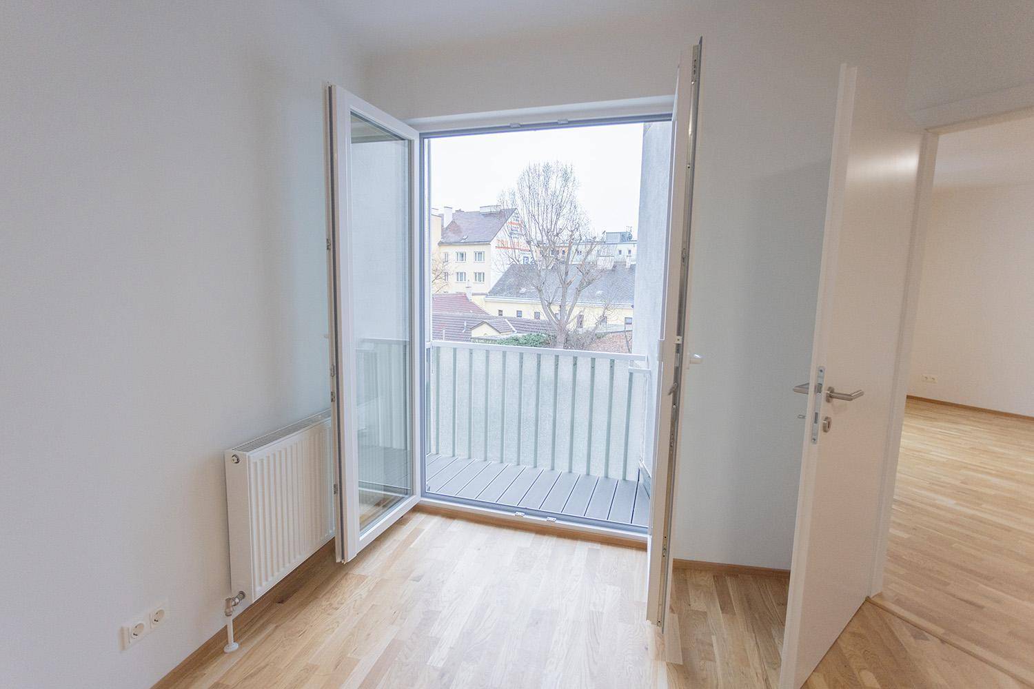 Heller Raum mit Holzboden und offener Balkontür, die einen Blick nach draußen freigibt.