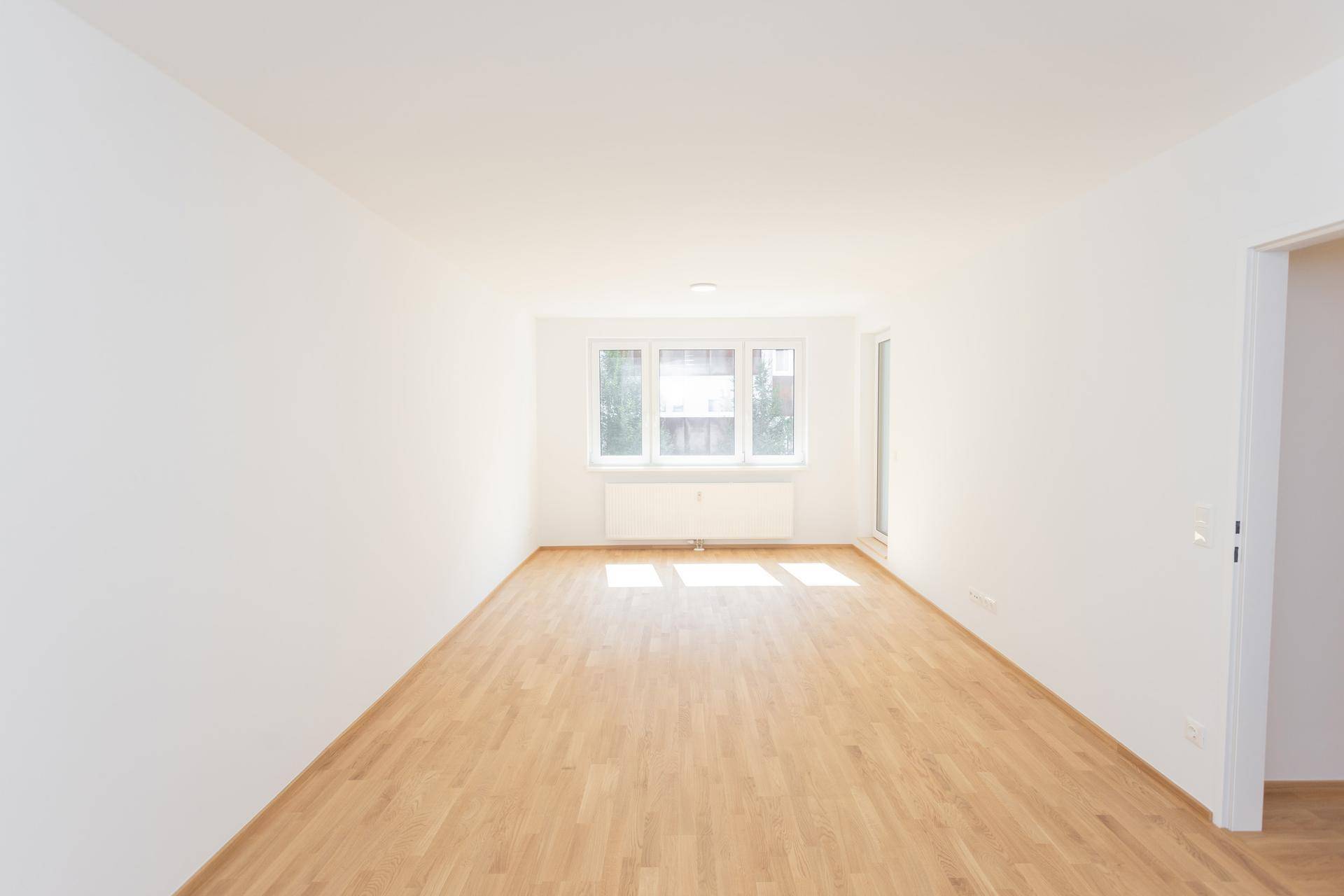 Geräumiges, unmöbliertes Zimmer mit hellem Holzparkett und großen Fenstern, die viel Tageslicht hereinlassen.