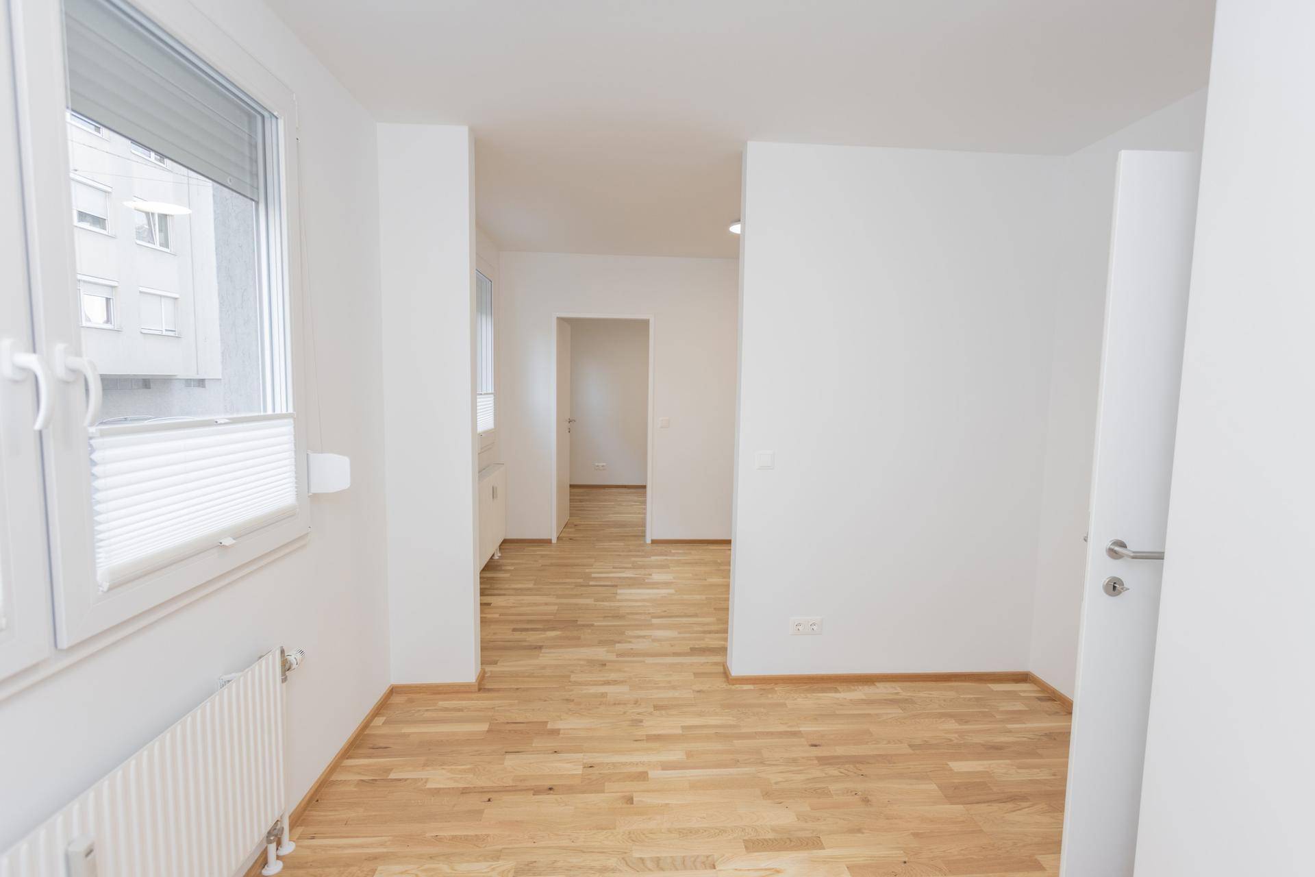 Leerer Raum mit Parkettboden und weißen Wänden, der den Blick in angrenzende Bereiche ermöglicht.