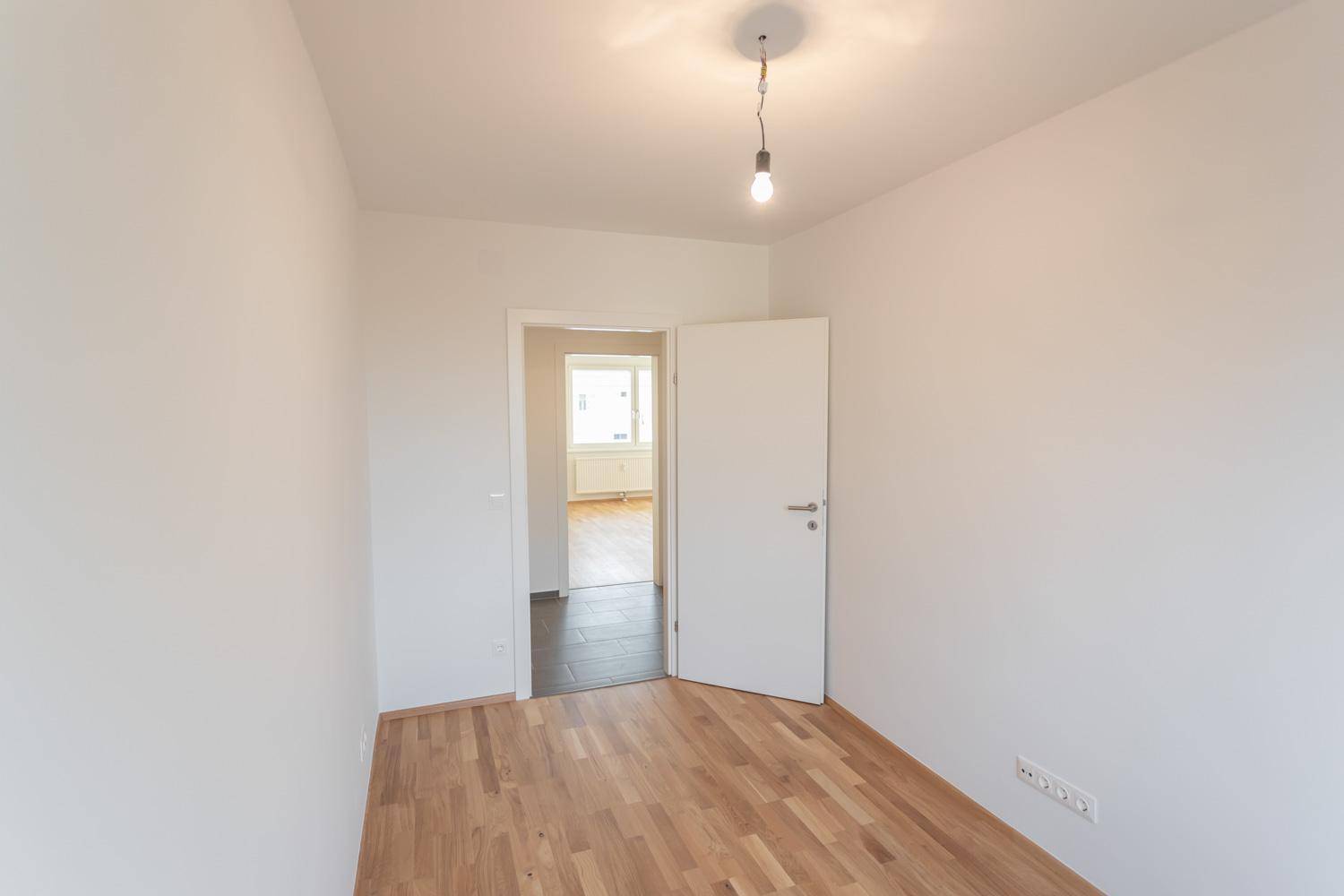 Leeres Zimmer mit Parkettboden und offener Tür zu einem weiteren Bereich.