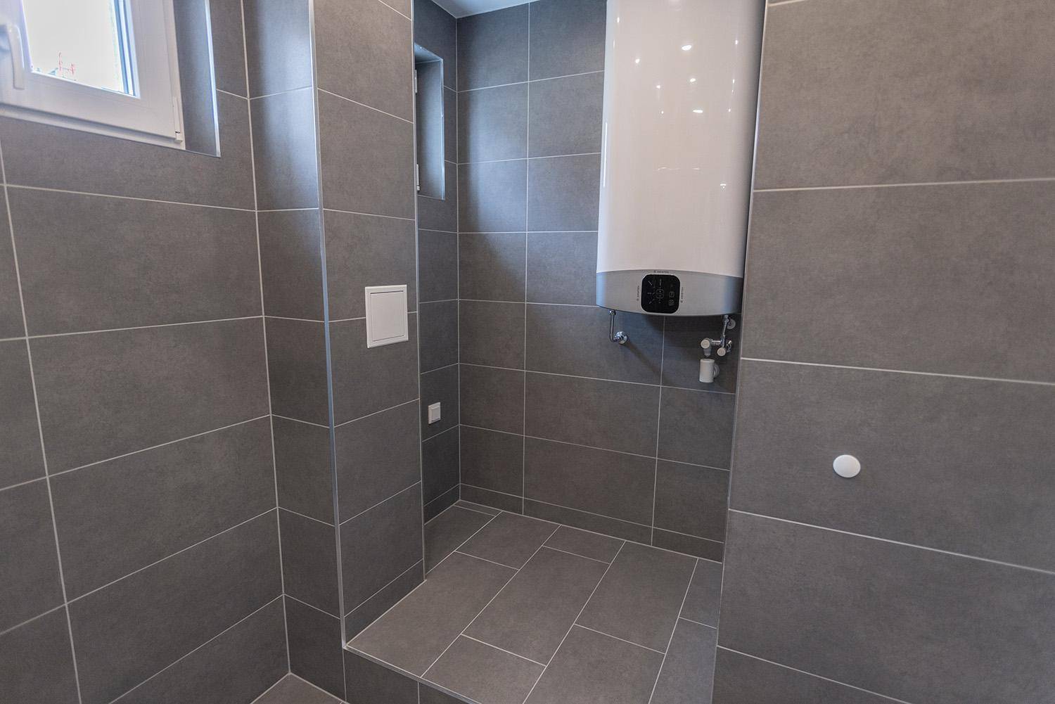 Modernes Badezimmer mit grauen Fliesen, Fenster und Warmwasserboiler, schlicht und funktional.