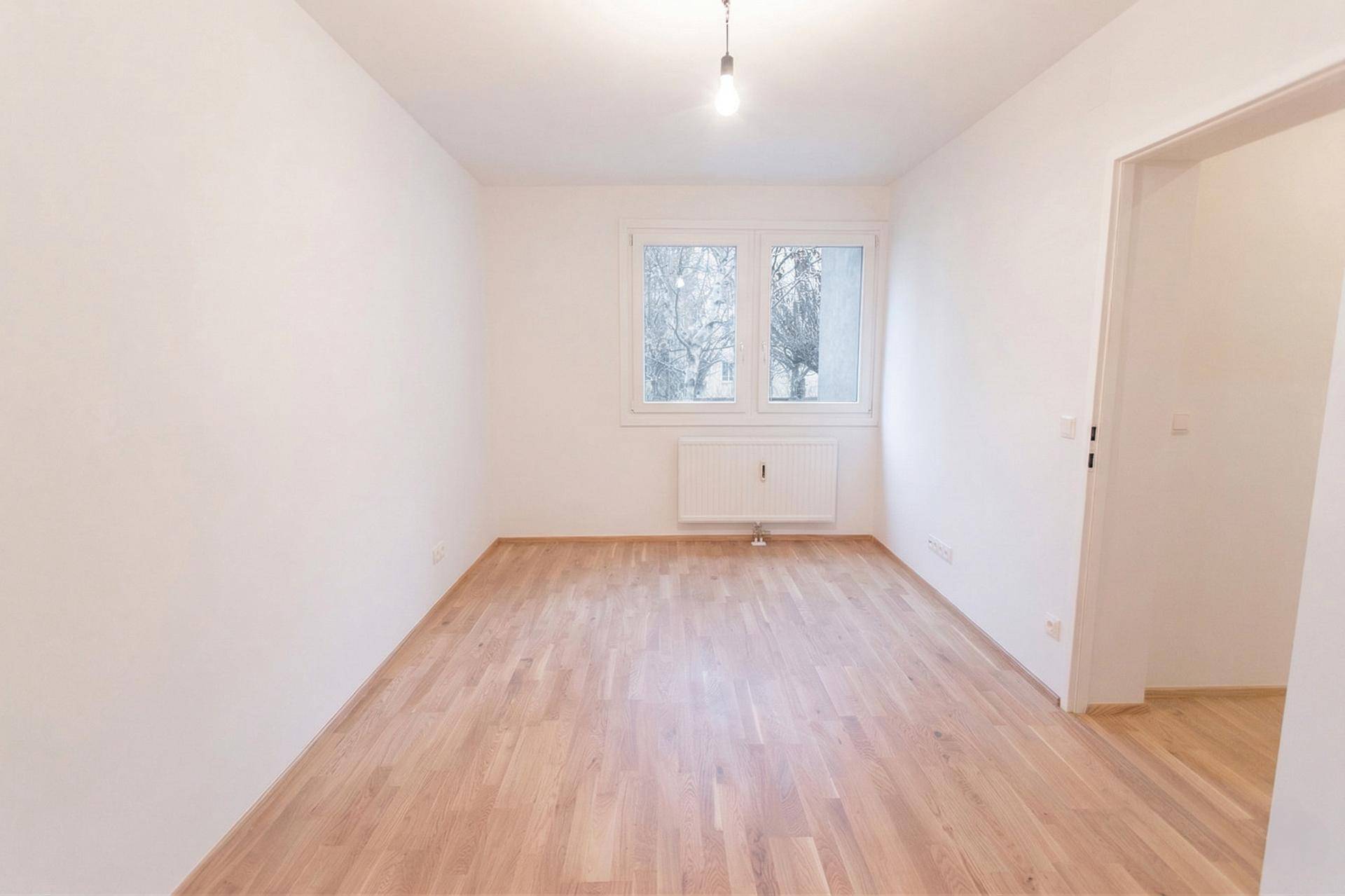 Heller, leerer Raum mit Parkettboden und einem Fenster, das viel Tageslicht hereinlässt.