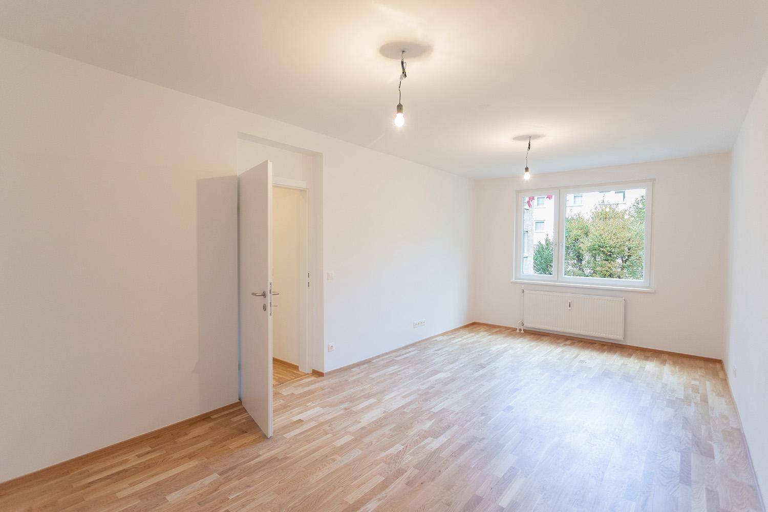 Helles, leeres Zimmer mit Holzboden und großem Fenster, bereit zur individuellen Gestaltung.