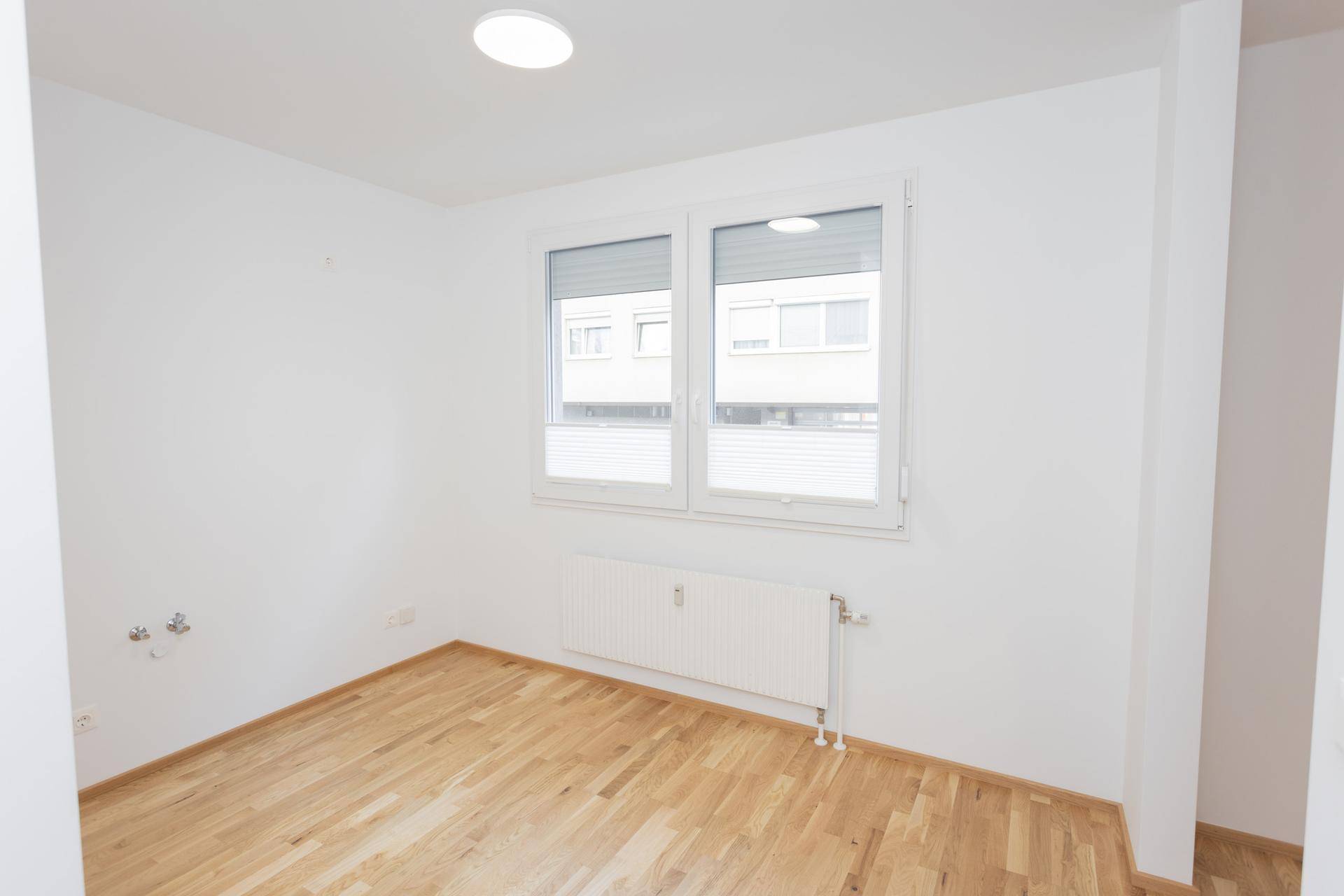 Leerer Raum mit hellem Parkettboden, Fenster mit Rollläden und Heizkörper, bereit zur Einrichtung.