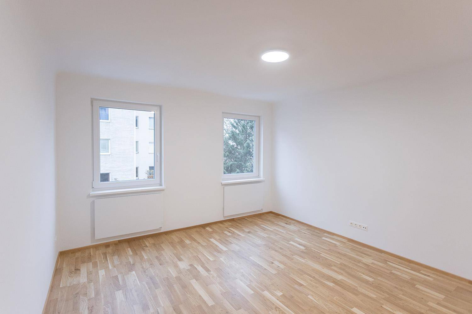 Helles Zimmer mit Holzboden und zwei Fenstern, die einen Blick ins Grüne bieten.