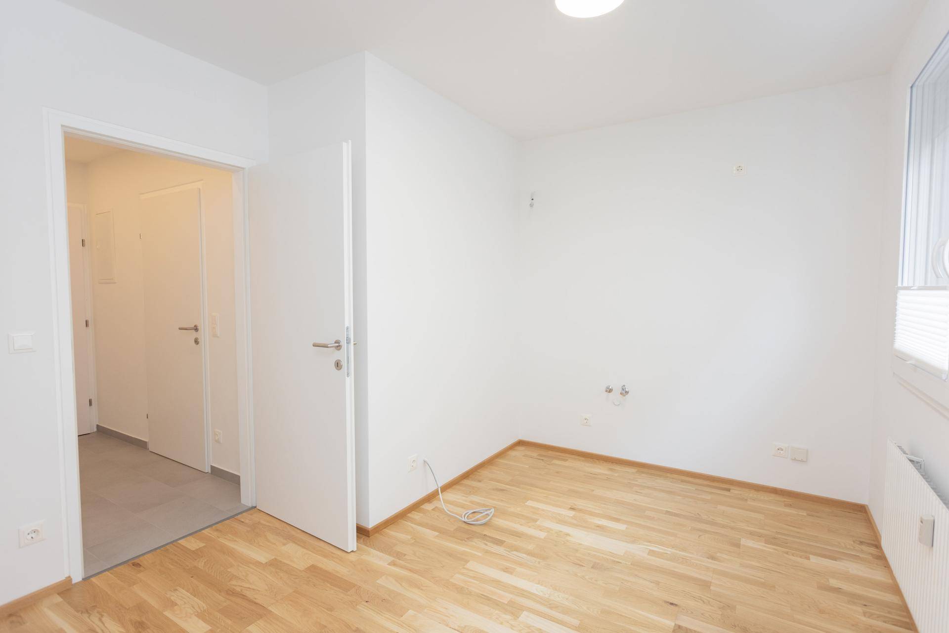 Leerer Raum mit Parkettboden und weiß gestrichenen Wänden, mit Blick in den Flur.