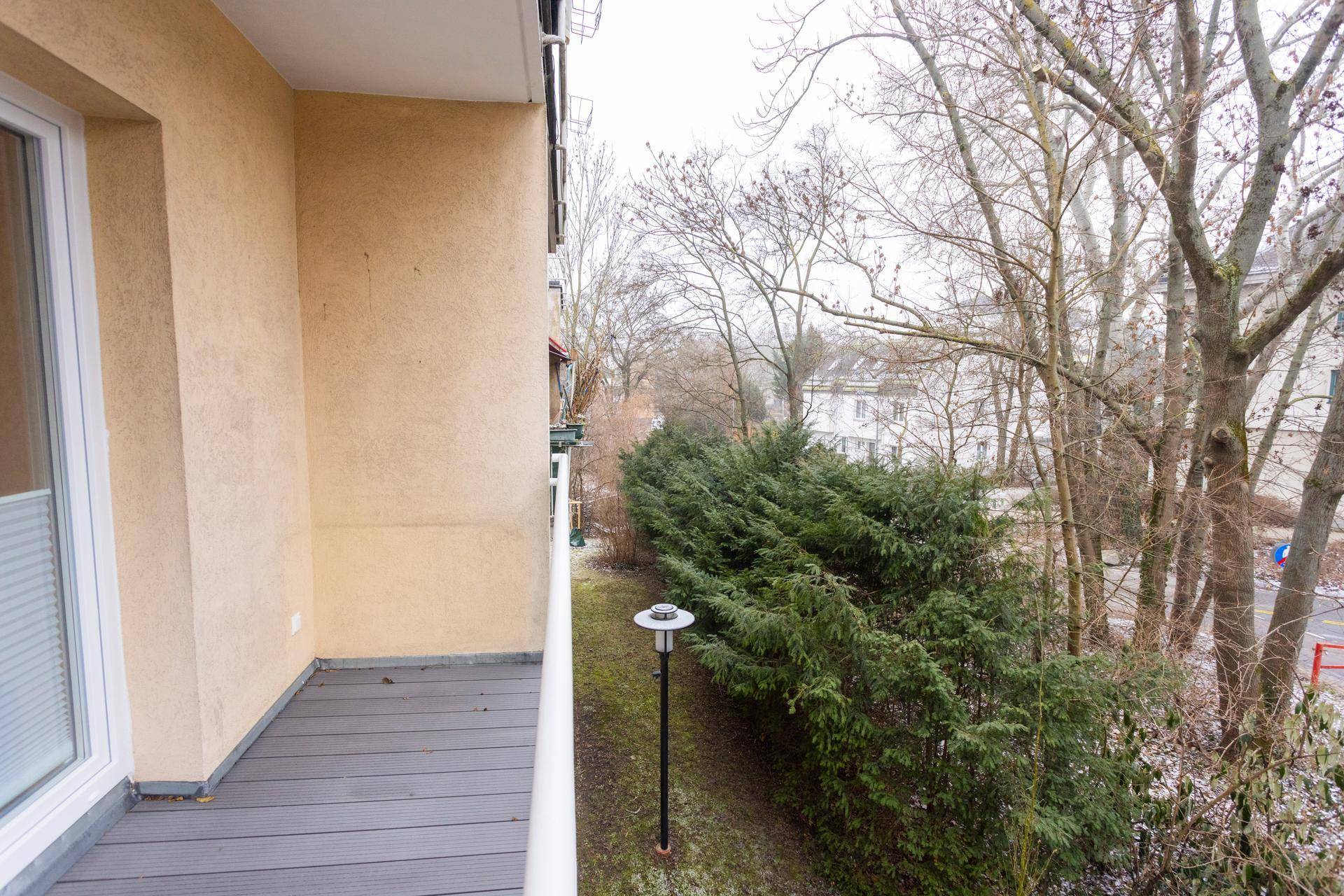Schmaler Balkon mit grauen Dielen und Blick auf eine Hecke und Bäume.
