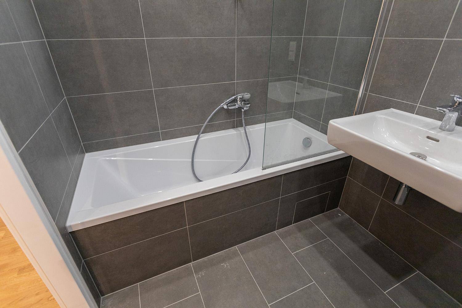 Modernes Badezimmer mit dunklen Fliesen, Badewanne mit Duschabtrennung und Waschtisch.