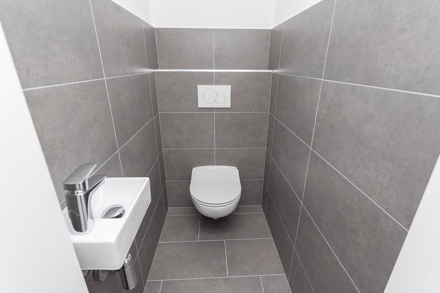 Separates WC mit Wand-WC, kleinem Waschbecken und grauen Fliesen, modern und sauber.