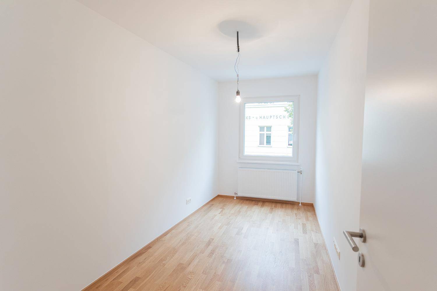 Leeres Zimmer mit hellem Holzboden, weiß gestrichenen Wänden und Fenster mit Ausblick.