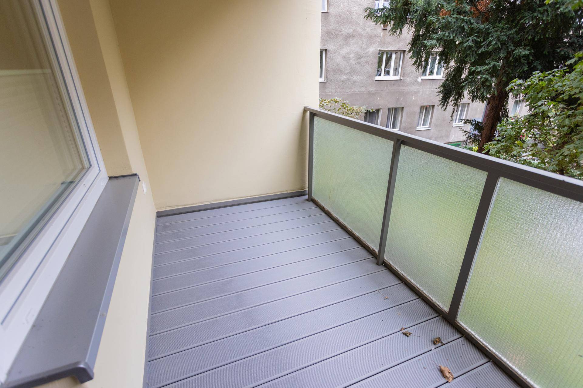 Balkon mit grauem Dielenboden und Blick auf eine grüne Fläche und angrenzende Gebäude.