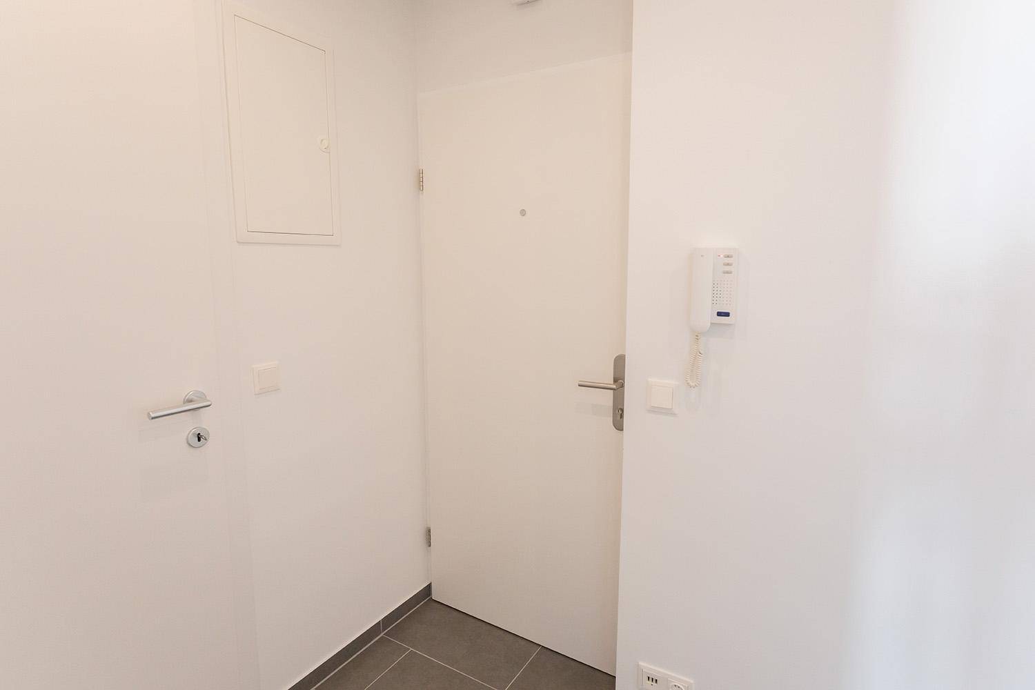 Eingangsbereich mit weißer Tür, Gegensprechanlage und grauen Fliesen, schlicht und funktional.