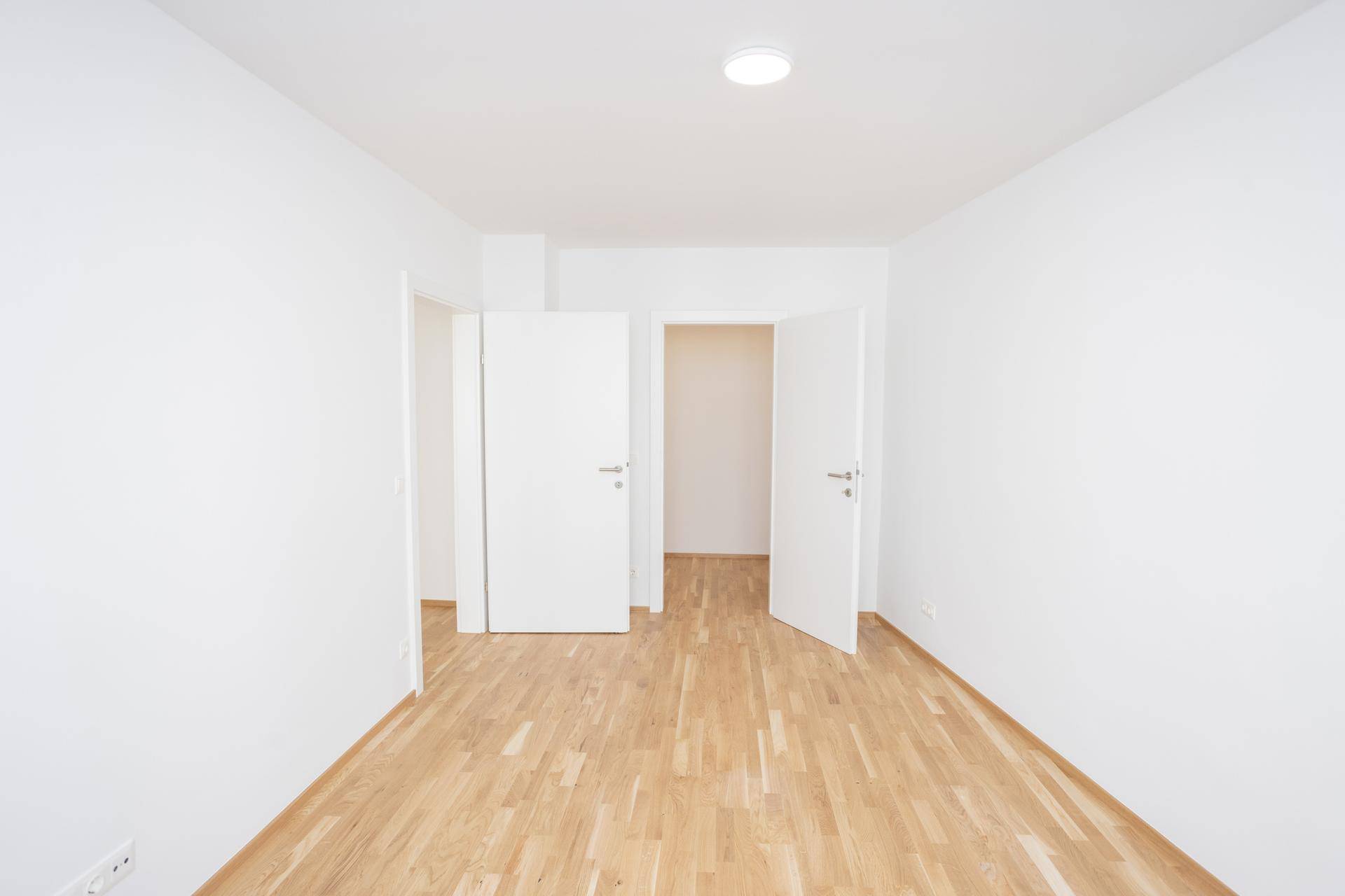 Leeres Zimmer mit hellem Parkettboden und zwei offenen Türen, das vielseitig nutzbar ist.