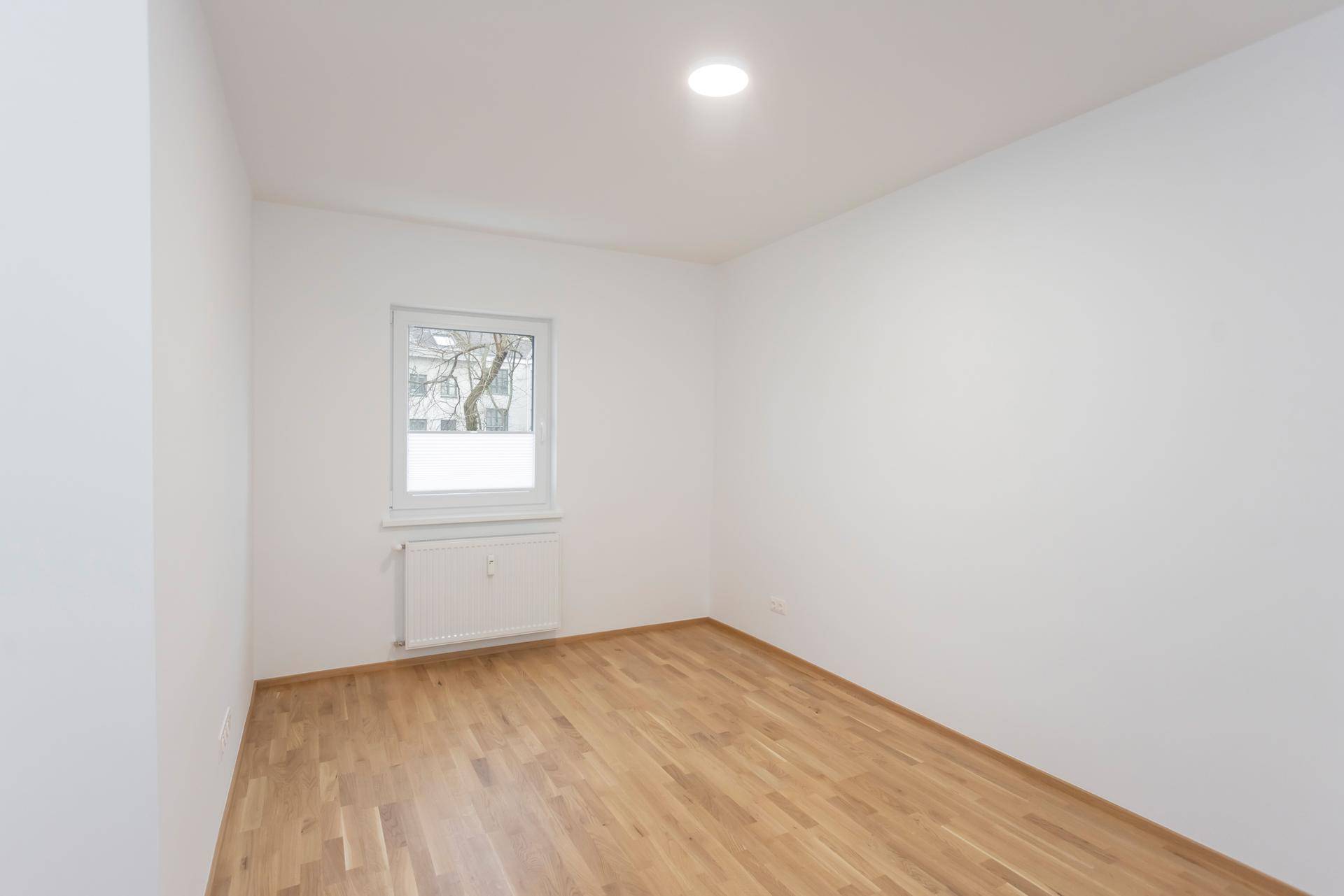 Helles Schlafzimmer mit Parkettboden und einem Fenster mit Blick ins Grüne.