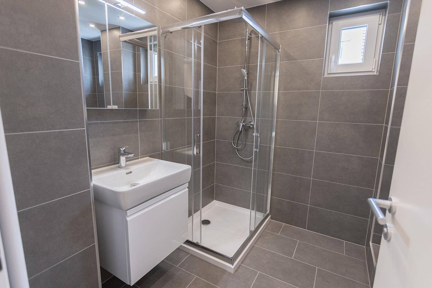 Modernes Badezimmer mit grauen Fliesen, Duschkabine und Waschtisch, funktional und sauber.