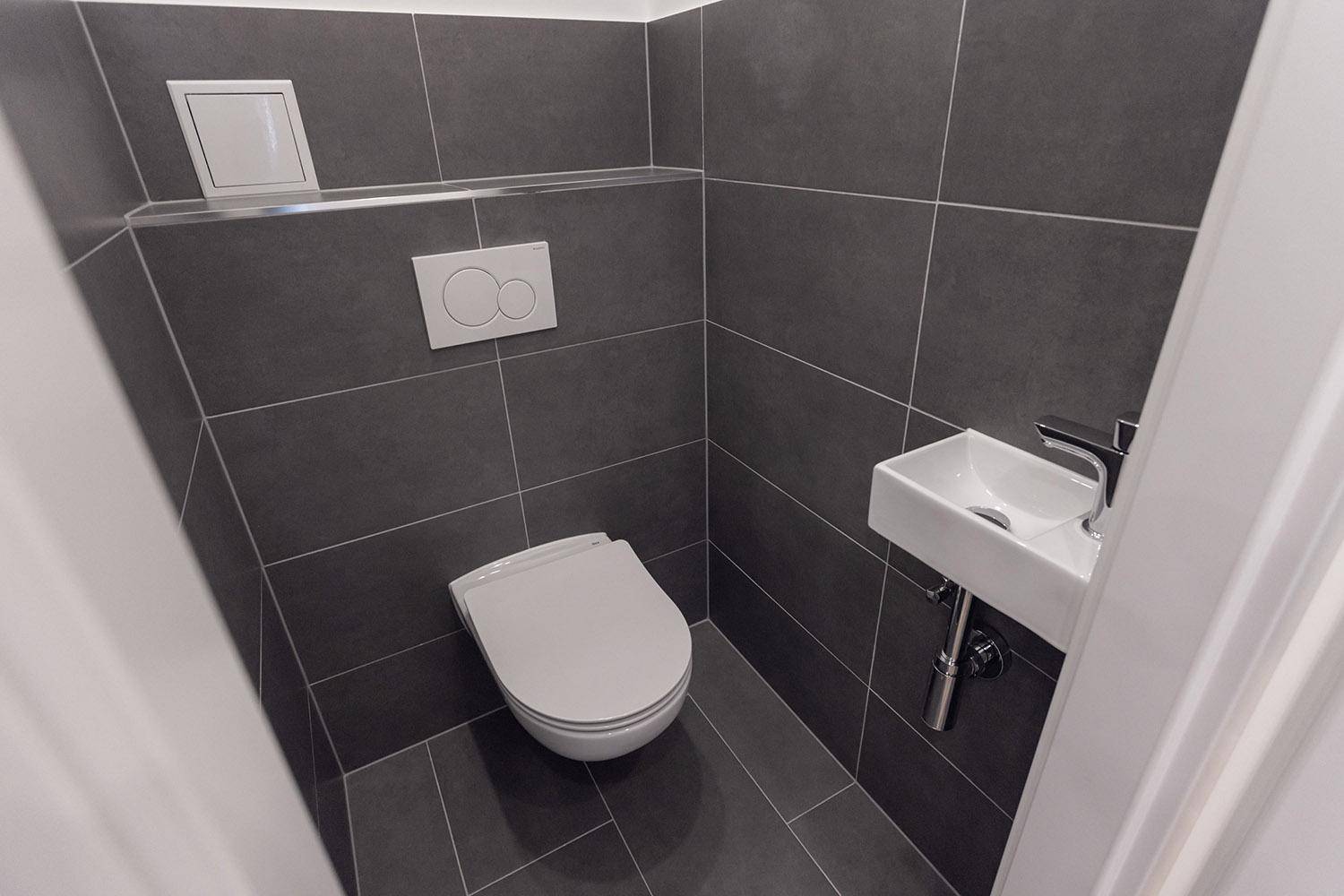 Modernes Gäste-WC mit dunklen Fliesen, wandhängendem WC und kleinem Waschbecken.