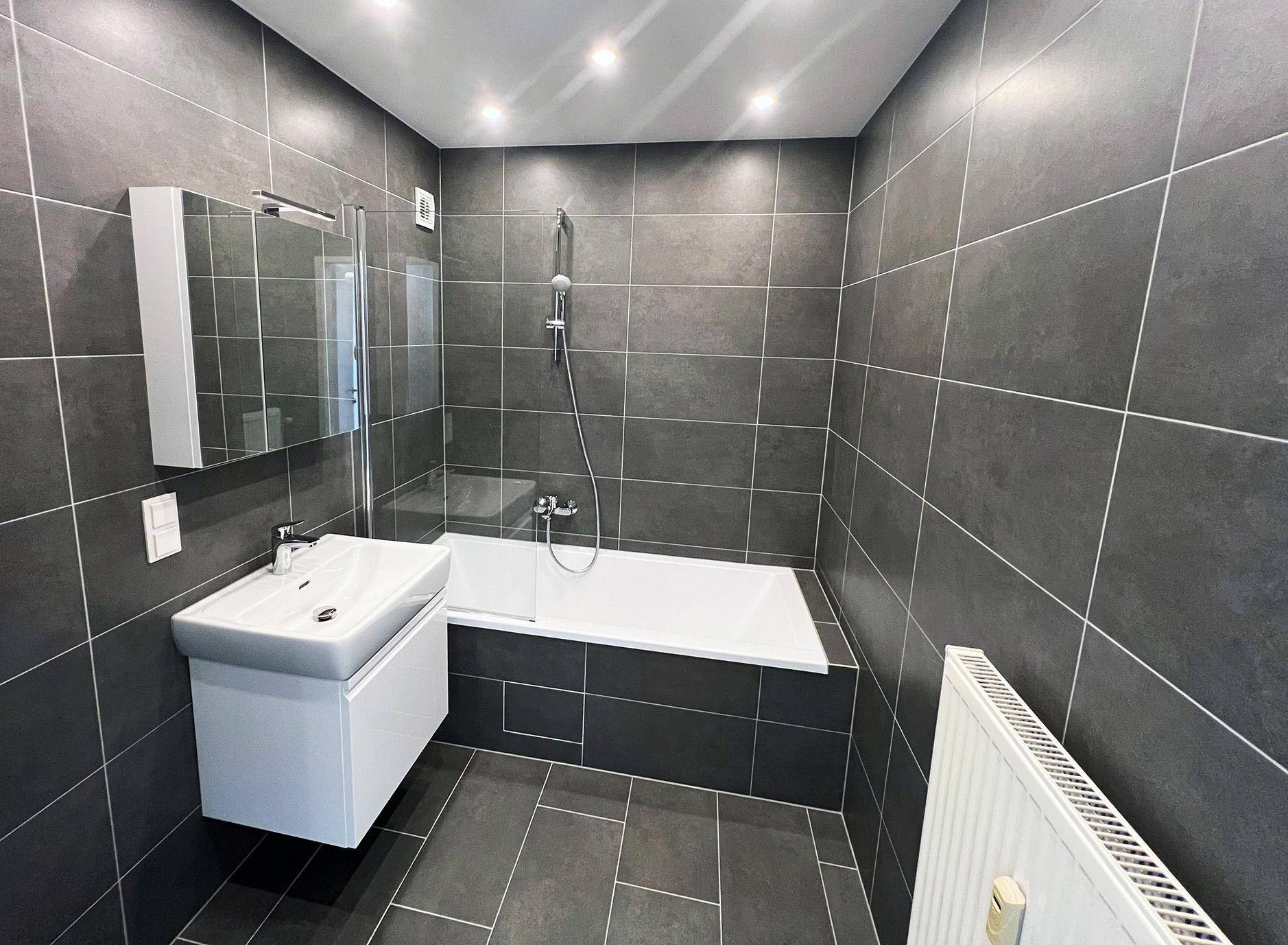Stilvolles Badezimmer mit dunklen Fliesen, Badewanne, Waschtisch und Heizkörper für Komfort.