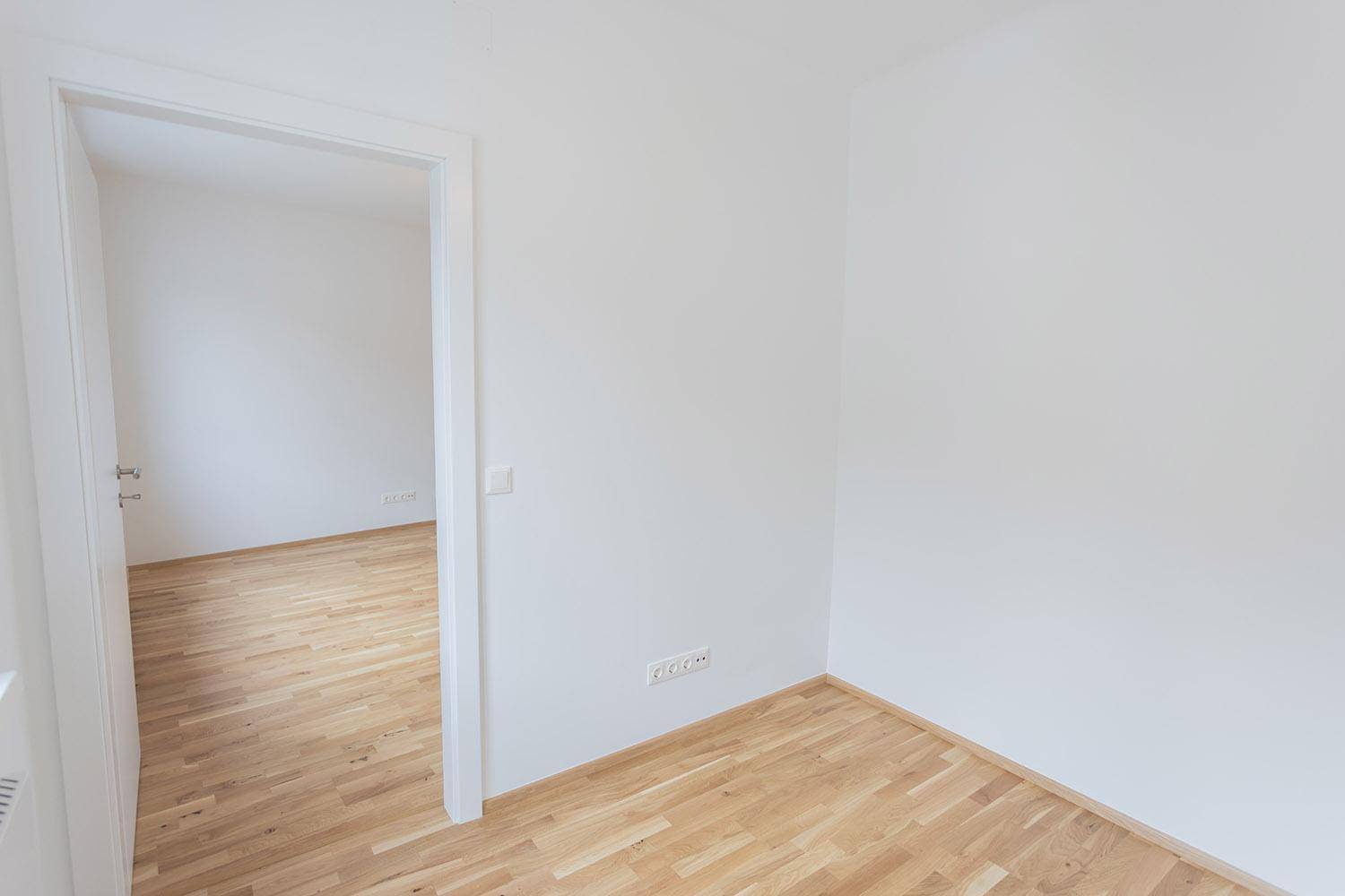 Leeres Zimmer mit Holzboden und weiß gestrichenen Wänden, das den Zugang zu einem weiteren Raum bietet.