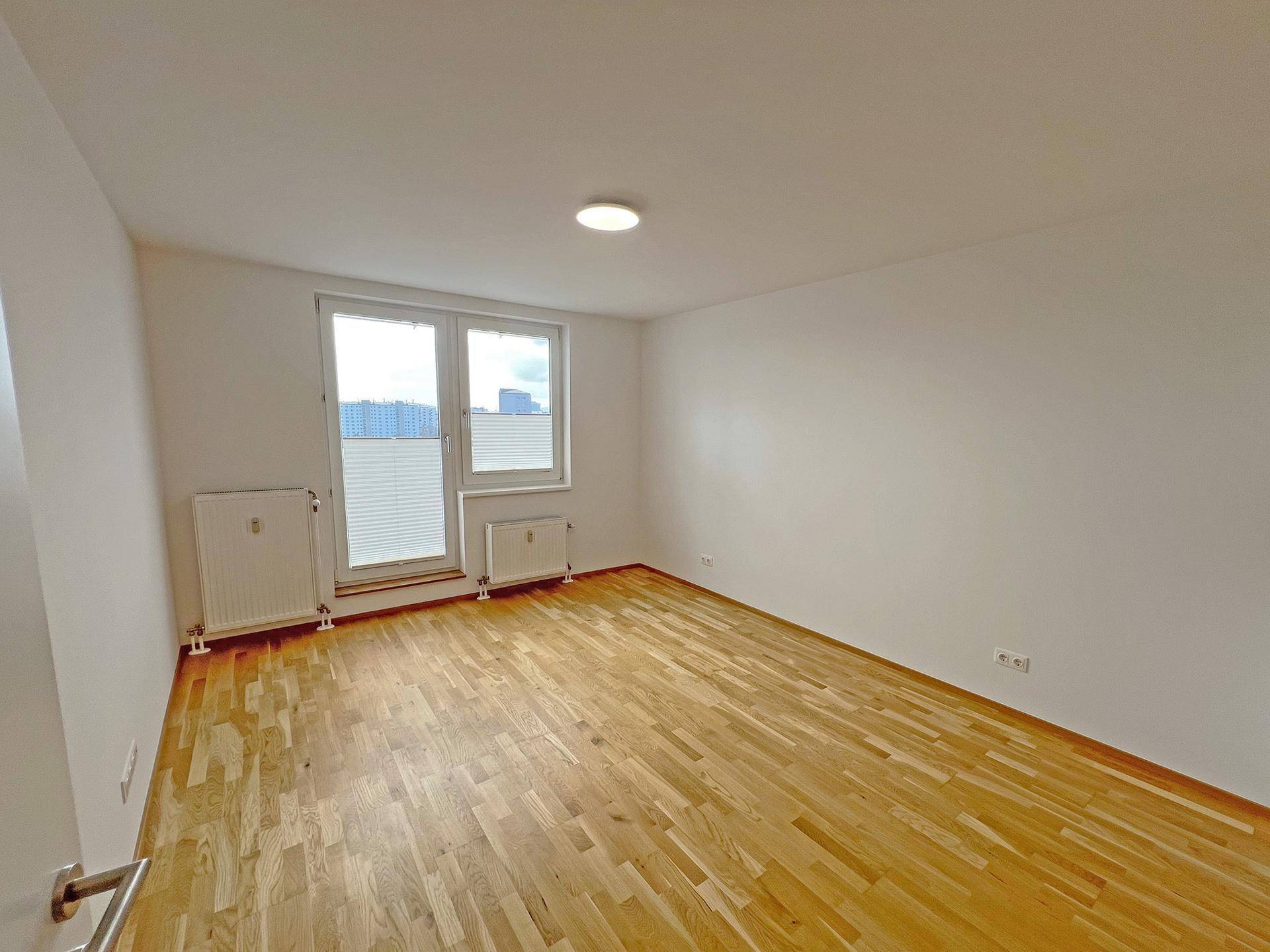 Helles, leeres Zimmer mit Parkettboden und Fenster mit Jalousien und Stadtblick.