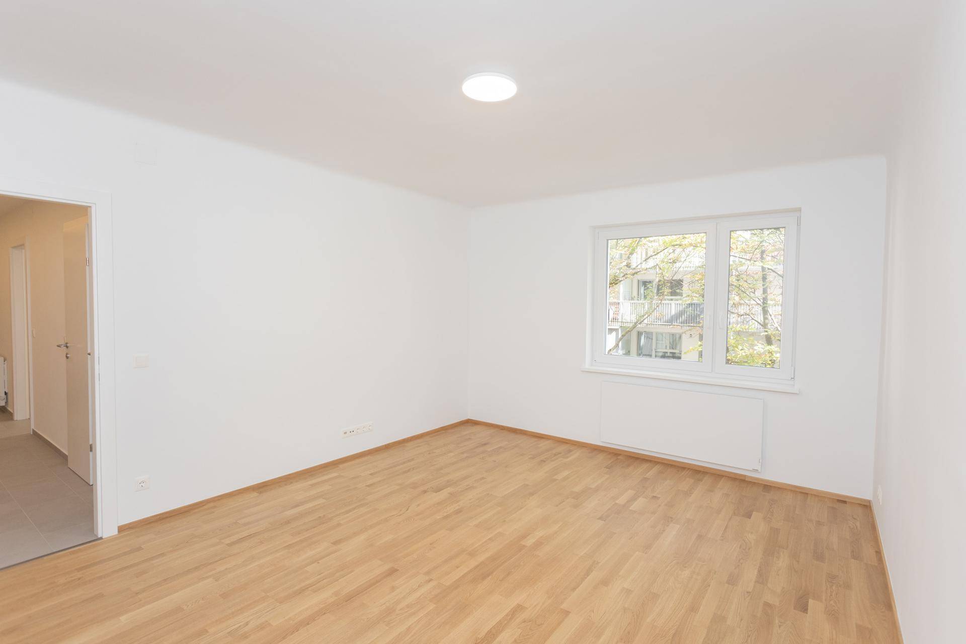 Helles, leeres Wohnzimmer mit Holzparkettboden und einem Fenster, das viel Tageslicht hereinlässt.