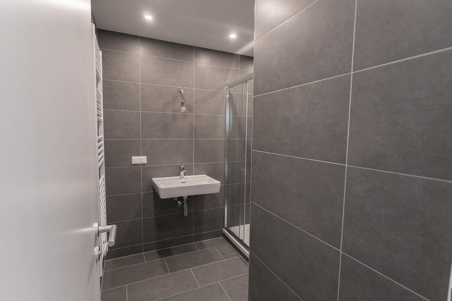 Modernes Badezimmer mit Waschbecken, Duschkabine und grauen Wand- und Bodenfliesen, hell beleuchtet.