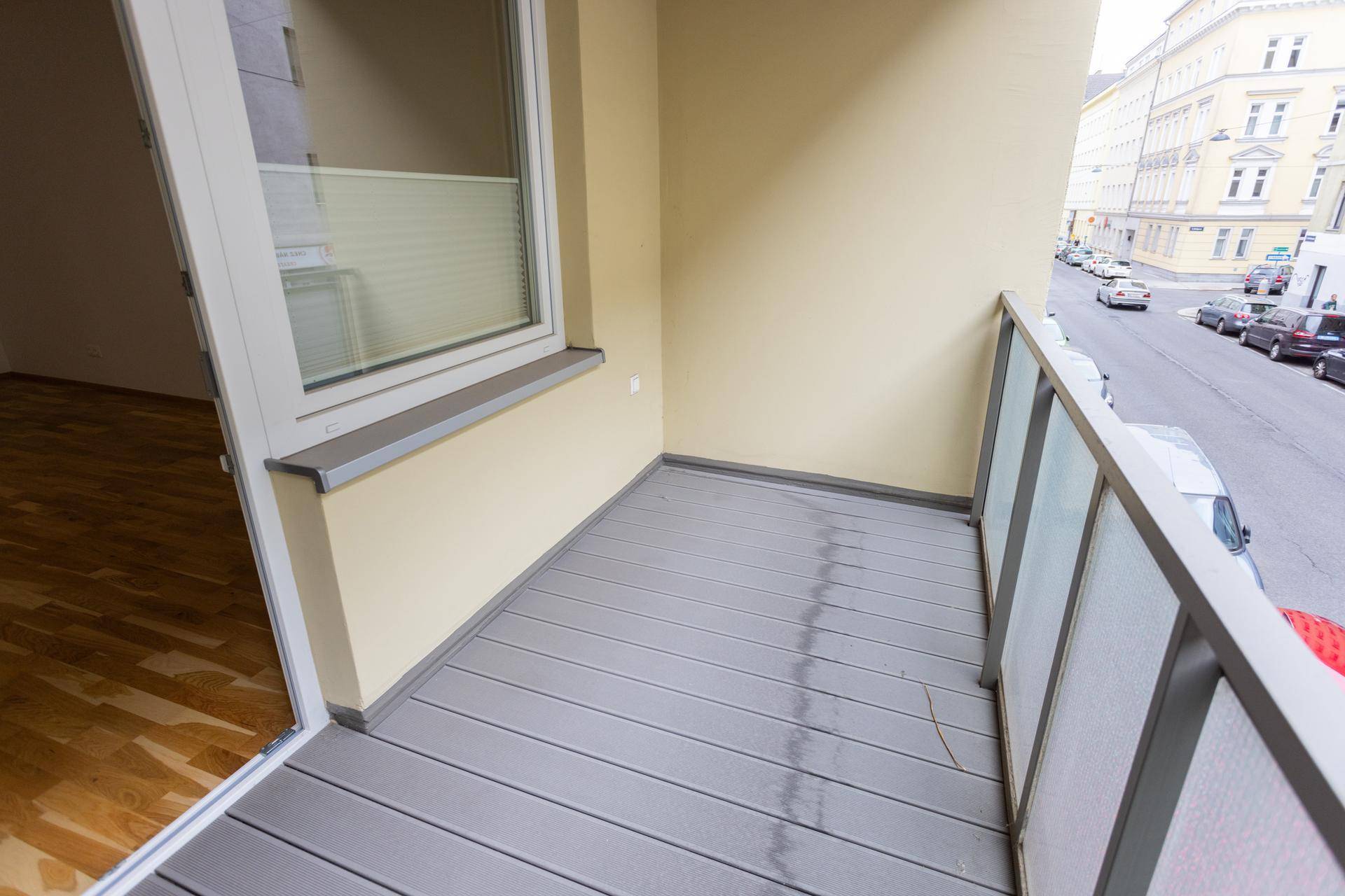 Balkon mit grauem Dielenboden und Glasgeländer, bietet Zugang zu einem Innenraum und Blick auf die Straße.