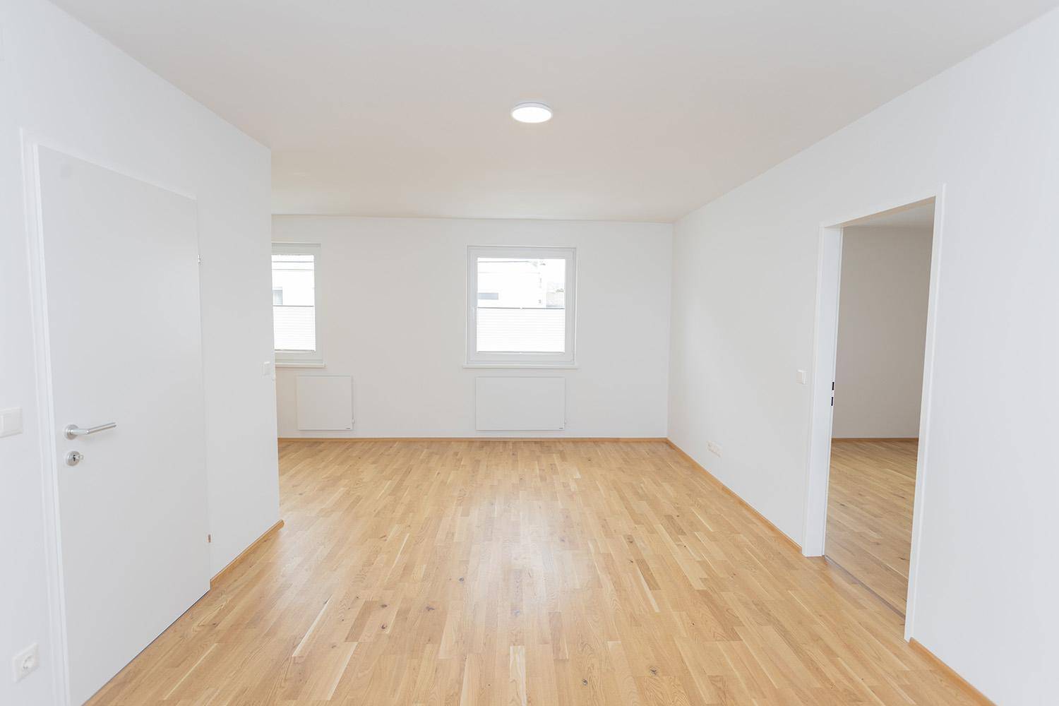 Geräumiges, leeres Wohnzimmer mit hellem Eichenparkett und zwei Fenstern, bereit zur Einrichtung.