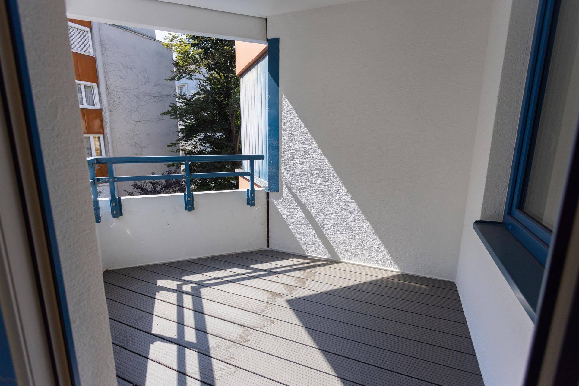 Sonniger Balkon mit modernem grauen Dielenboden und blauer Brüstung, umgeben von Bäumen und Gebäuden.
