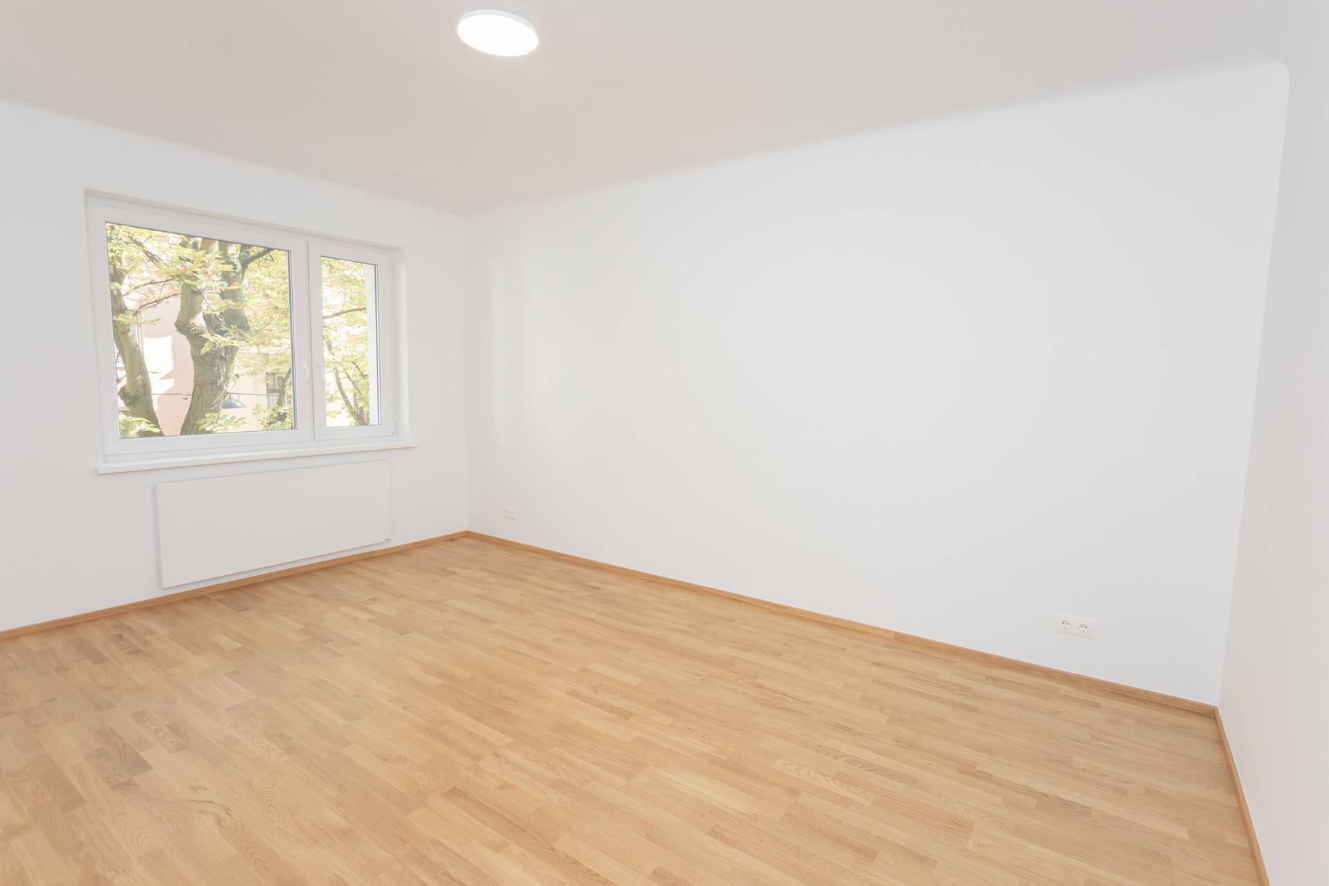 Helles, leeres Wohnzimmer mit Holzparkettboden und einem großen Fenster mit Blick ins Grüne.