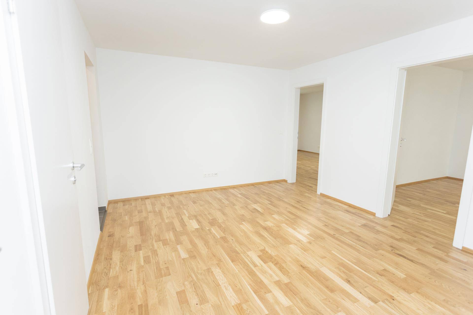 Leerer Raum mit hellem Parkettboden und Zugang zu weiteren Zimmern der Wohnung.