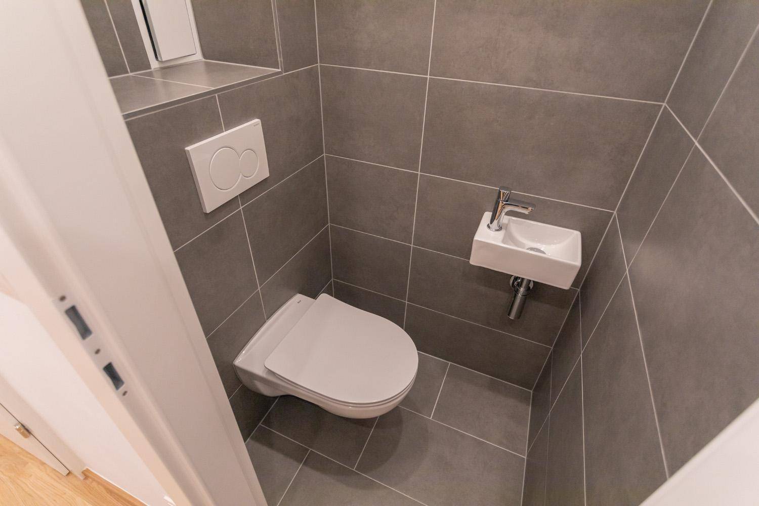 Modernes Gäste-WC mit grauen Fliesen, wandhängendem WC und kleinem Waschbecken.