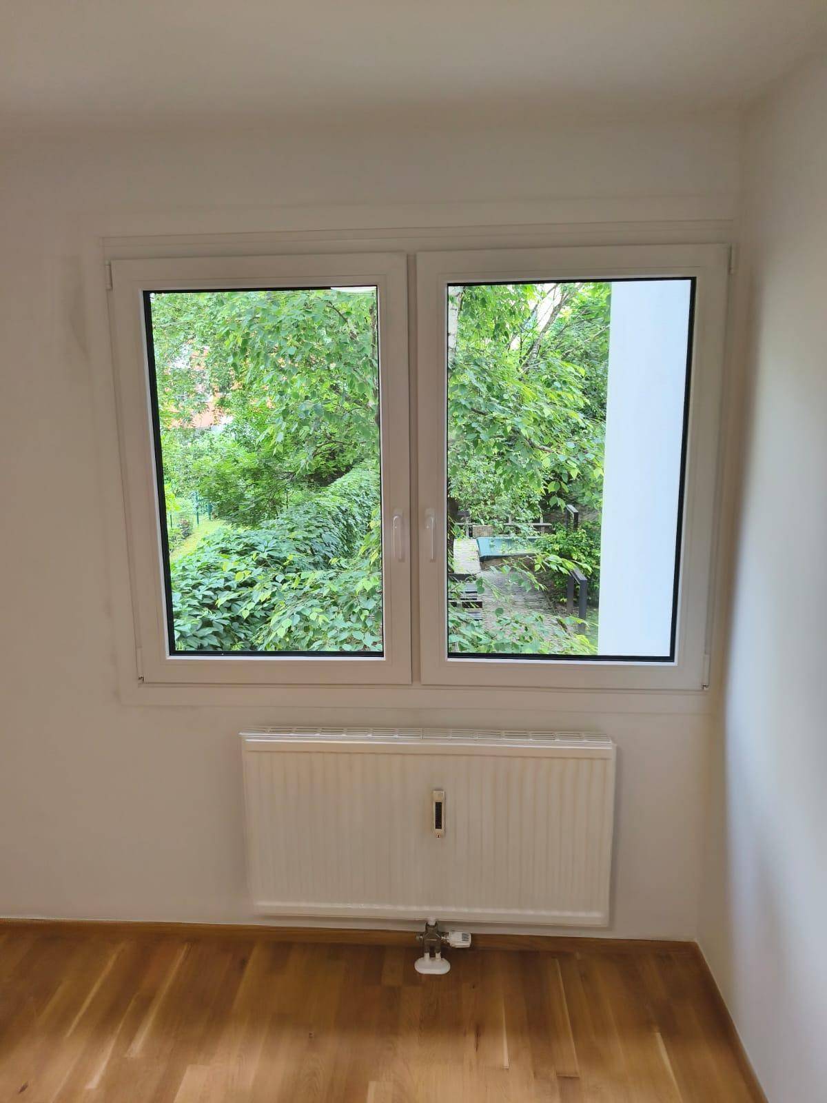 Heller Raum mit Parkettboden, weißem Fenster und Blick ins Grüne.