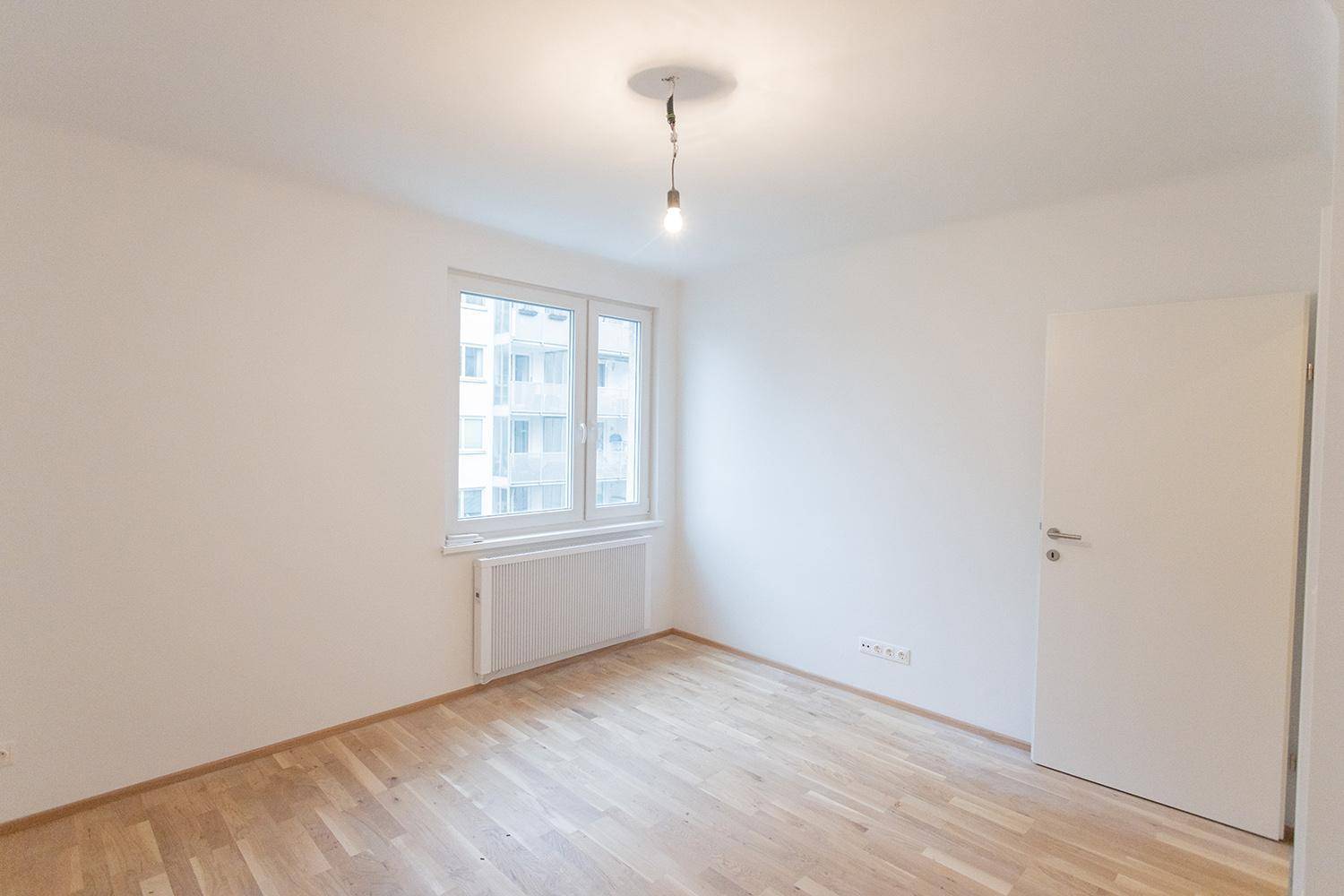 Geräumiges Zimmer mit Parkettboden, Fenster und weißer Tür, bietet viel Tageslicht.