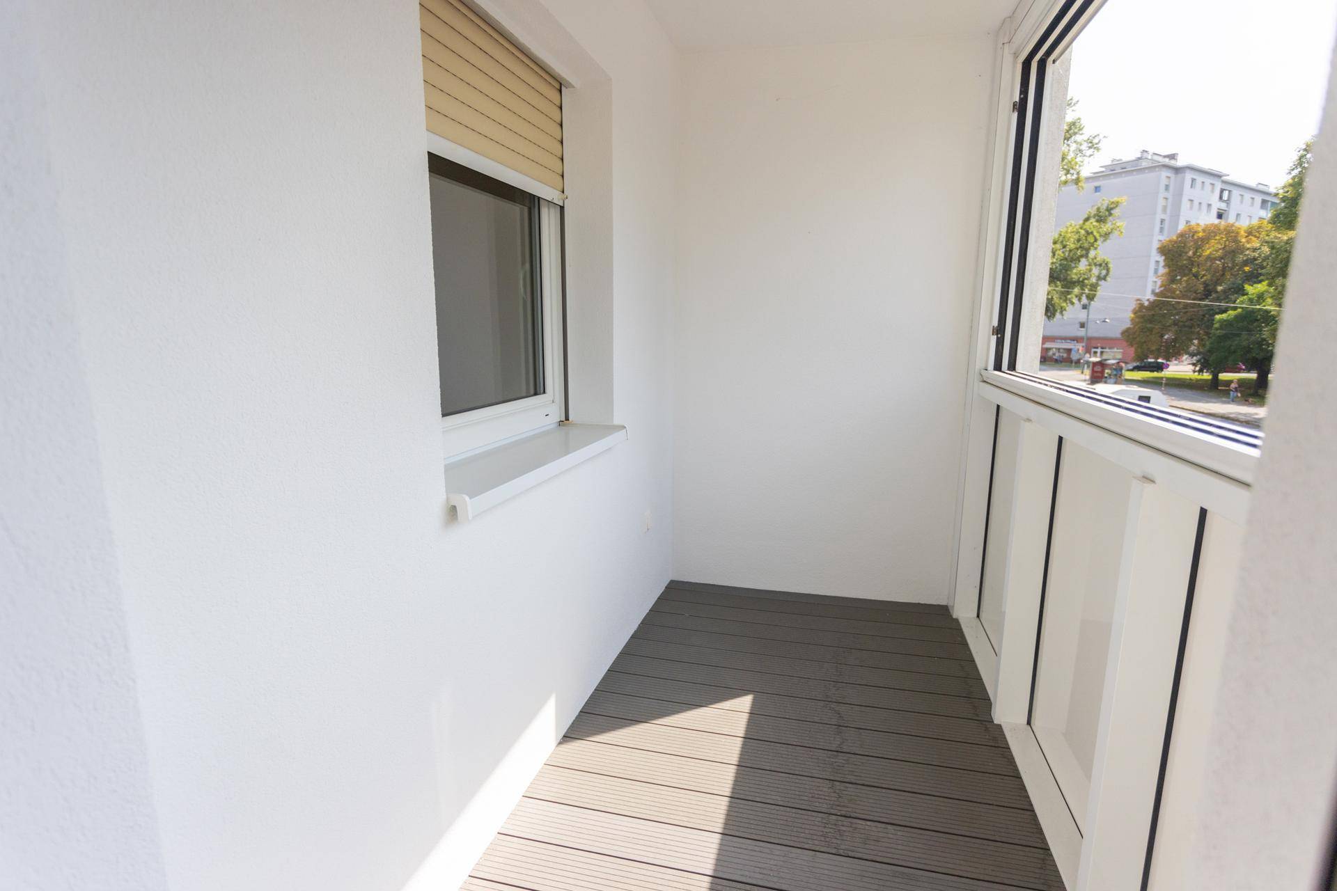 Heller Balkon mit Holzdielenboden und einem Fenster, das viel Tageslicht hereinlässt.