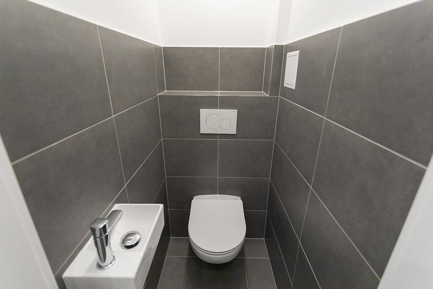 Separate Toilette mit Wand-WC, kleinem Waschbecken und grauen Fliesen.