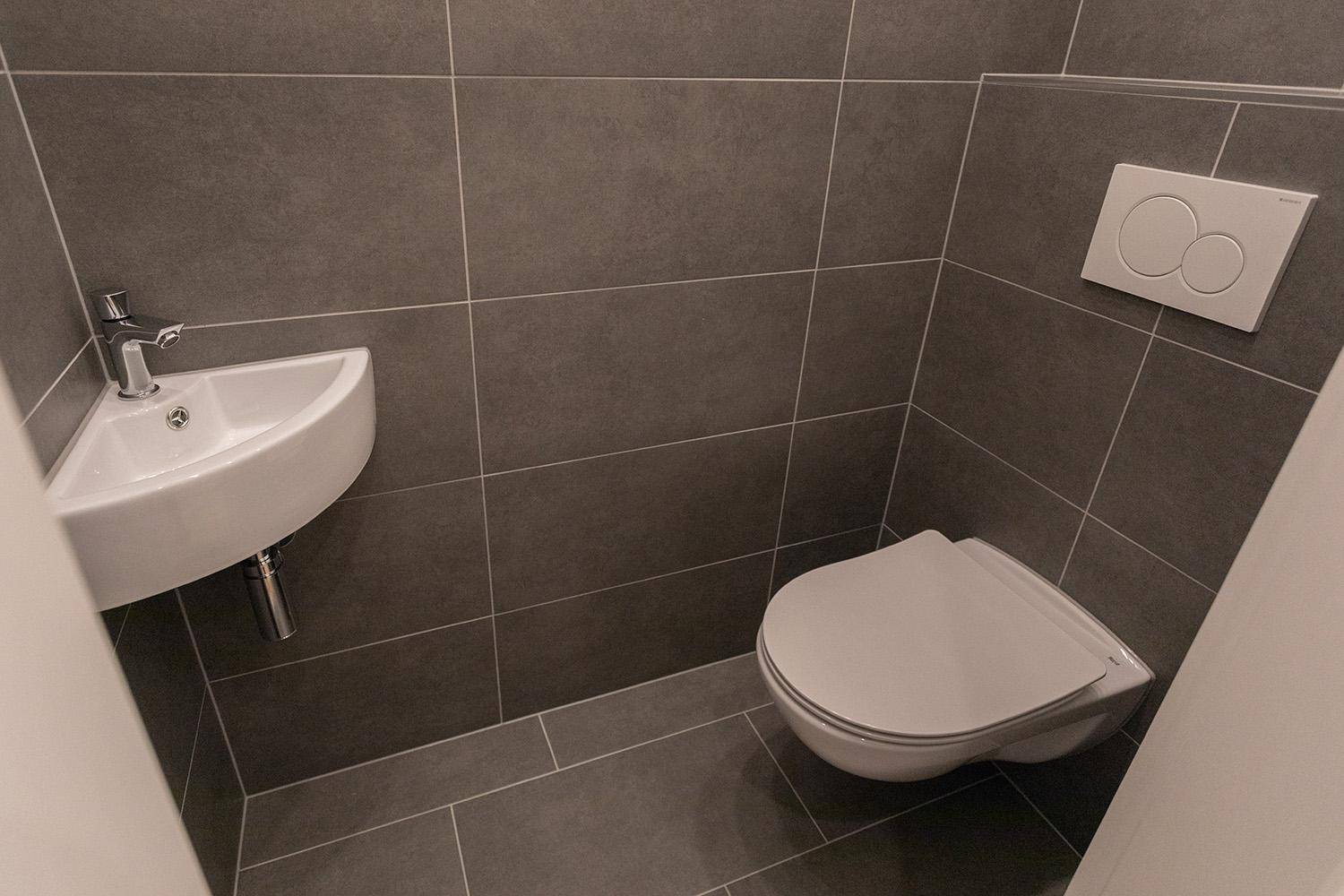 Modernes Gäste-WC mit grauen Fliesen, wandhängendem WC und kleinem Eckwaschbecken.