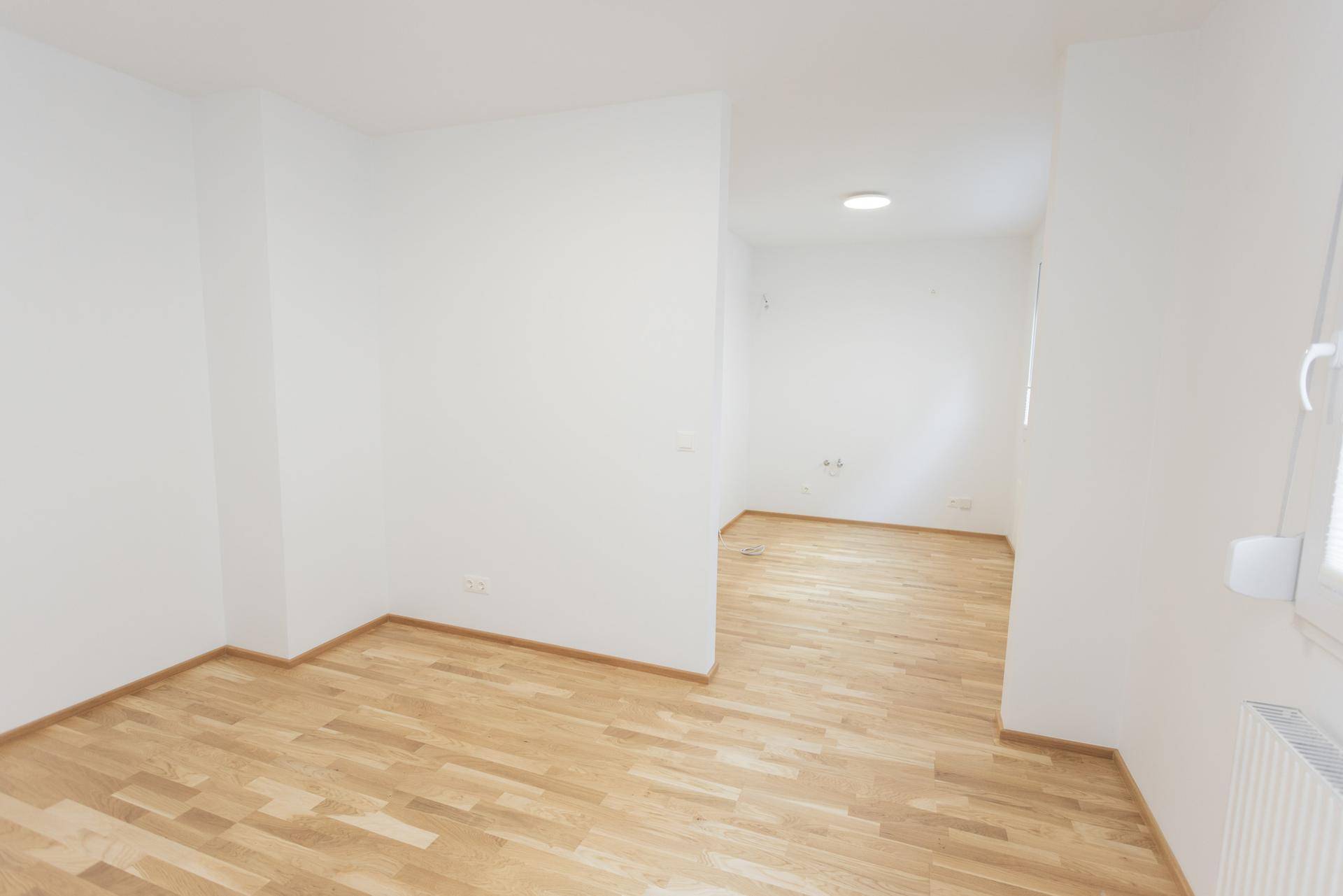 Leerer Raum mit Parkettboden und weißen Wänden, der den Blick in angrenzende Bereiche ermöglicht.