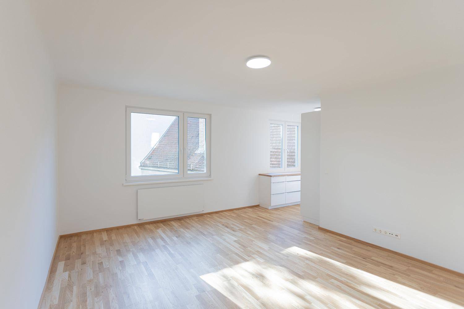 Heller, leerer Raum mit Holzboden und Fensterblick auf umliegende Dächer und Gebäude.